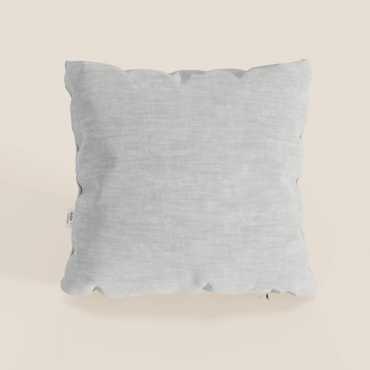 Un cuscino Sofia quadrato grigio chiaro di divitt.sbs in morbido velluto nuvolato Easy Clean T35, antimacchia e impermeabile, poggia su un fondo crema chiaro; è morbido, leggermente gonfio, con un'etichetta in tessuto bianco sul bordo sinistro.