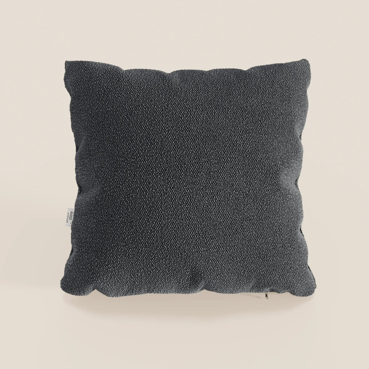 Un cuscino Lambada quadrato grigio antracite di divitt.sbs in tessuto smacchiabile e impermeabile tipo cotone naturale si trova in posizione verticale su uno sfondo beige chiaro con pieghe morbide e una piccola etichetta bianca sulla sinistra.