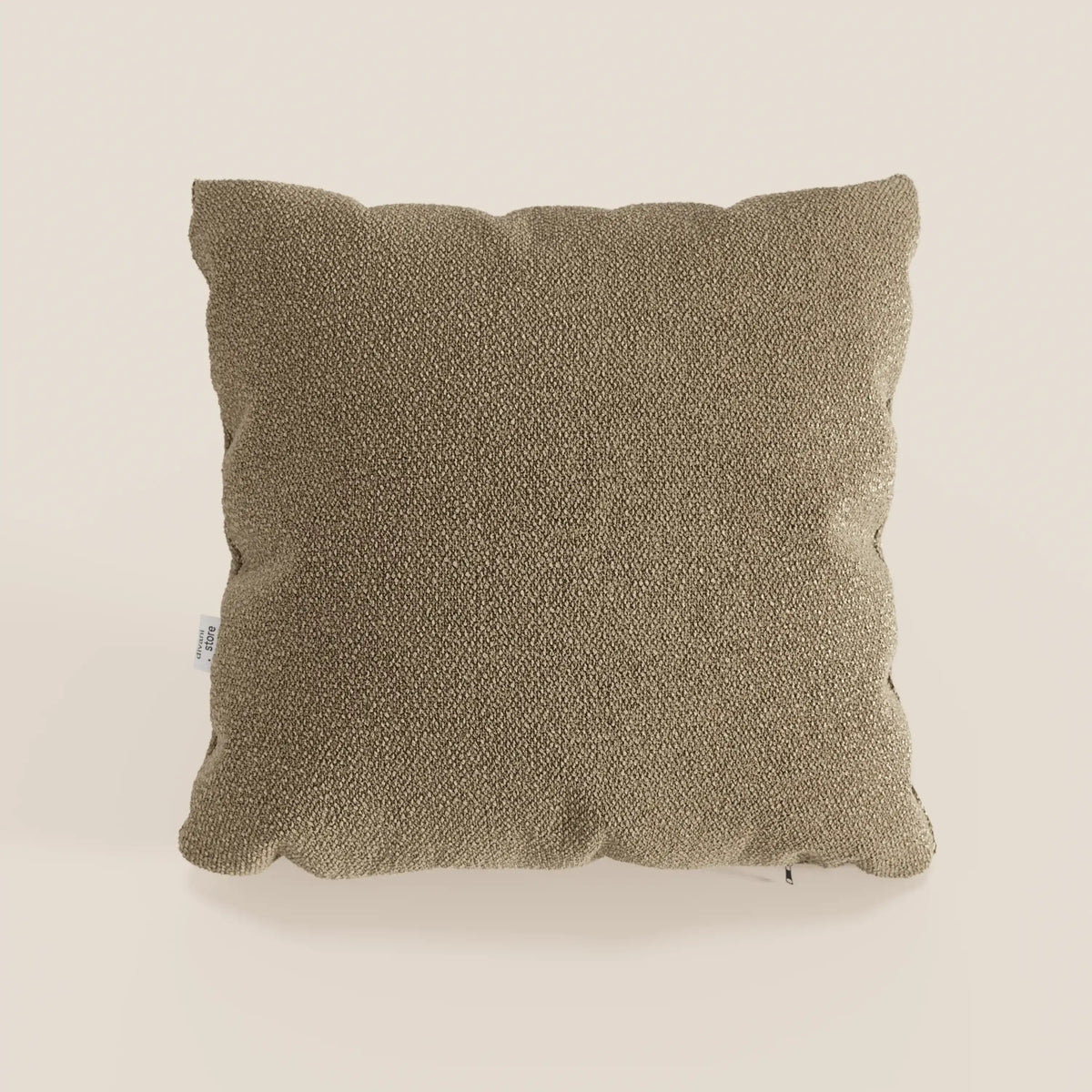 Un cuscino Lambada marrone di divitt.sbs, quadrato e strutturato, realizzato in tessuto T33 di cotone naturale, facile da pulire e impermeabile, è centrato su uno sfondo beige semplice. Sul lato sinistro è visibile una piccola etichetta bianca. L'illuminazione è uniforme e neutra.