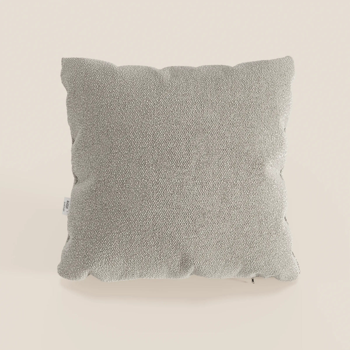 Cuscino Lambada quadrato grigio chiaro di divitt.sbs in tessuto T33 lavabile e impermeabile, simile al cotone, con una texture sottile, su uno sfondo bianco sporco. Una piccola etichetta bianca compare sul bordo sinistro del cuscino.