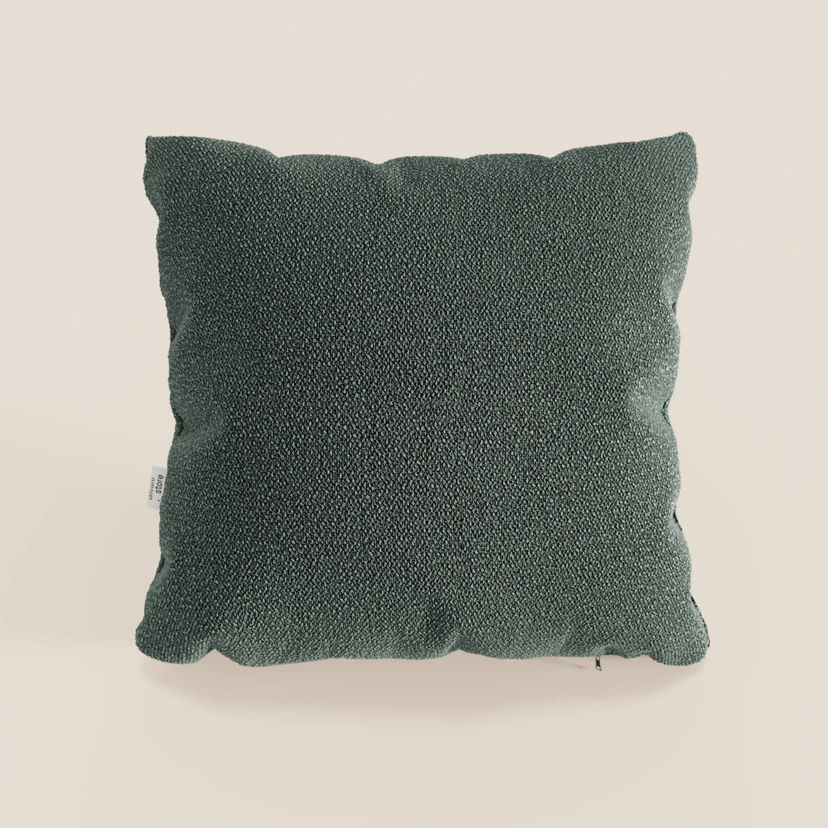 Il cuscino Lambada di divitt.sbs, di forma quadrata e di colore verde scuro, realizzato in tessuto smacchiabile e impermeabile effetto cotone naturale, si presenta con bordi leggermente arrotondati e una piccola etichetta bianca a sinistra su uno sfondo beige chiaro.