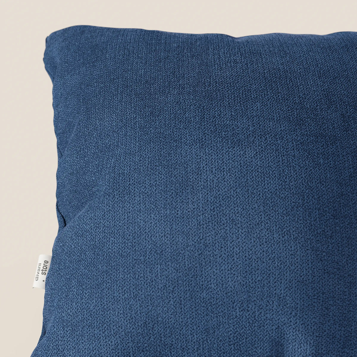 Primo piano del cuscino Destiny 45x45 di divitt.sbs in T02 blu, caratterizzato da un morbido tessuto impermeabile. Il cuscino è parzialmente rappresentato con una piccola etichetta in tessuto bianco con testo nero sulla cucitura sinistra, su uno sfondo bianco sporco.