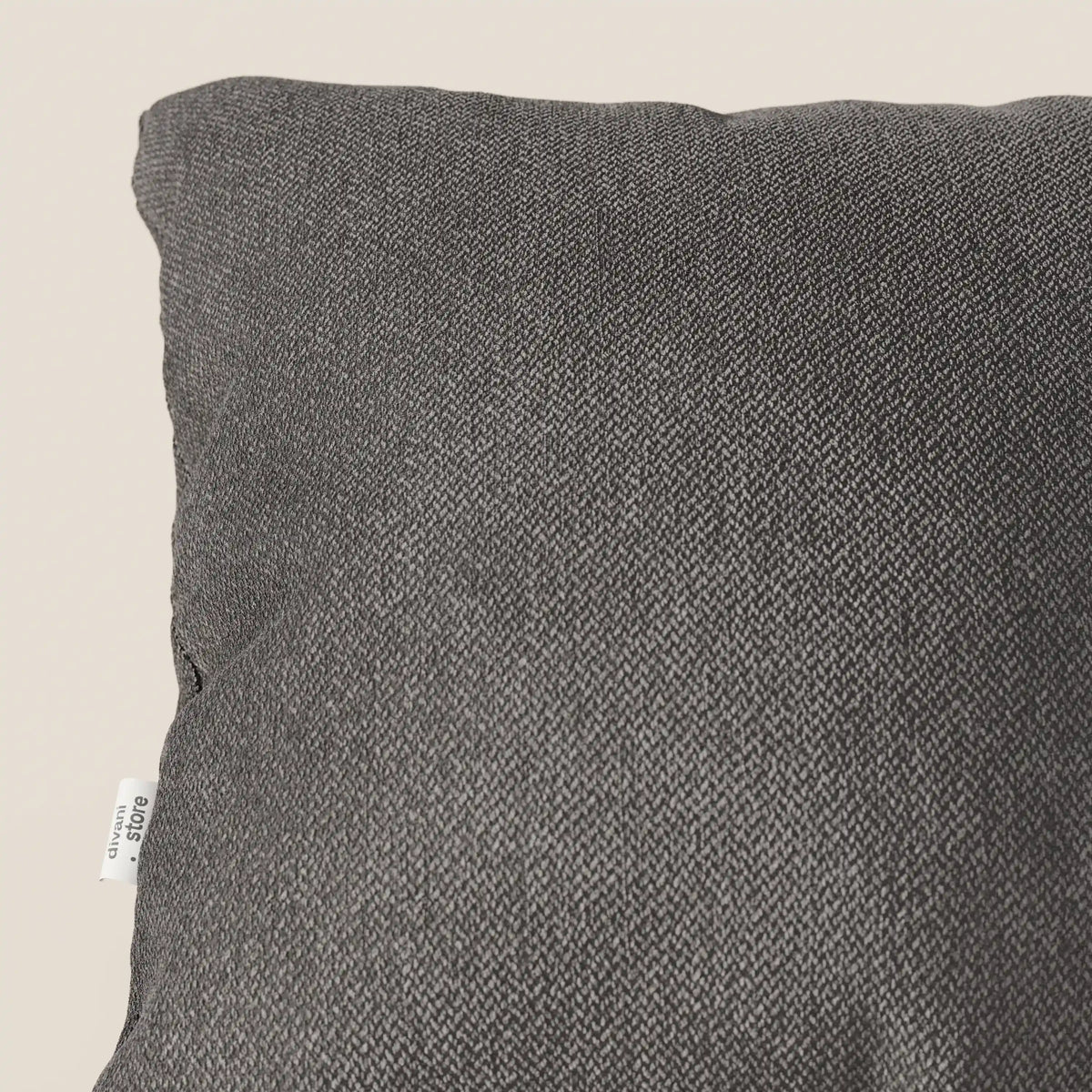Primo piano dell'angolo superiore sinistro di un cuscino Sogno di divitt.sbs in tessuto morbido SMACCHIABILE T05 antracite, su uno sfondo beige chiaro. Una piccola etichetta bianca con la scritta nera "&C URBAN" è cucita nella cucitura laterale di questo elegante cuscino.