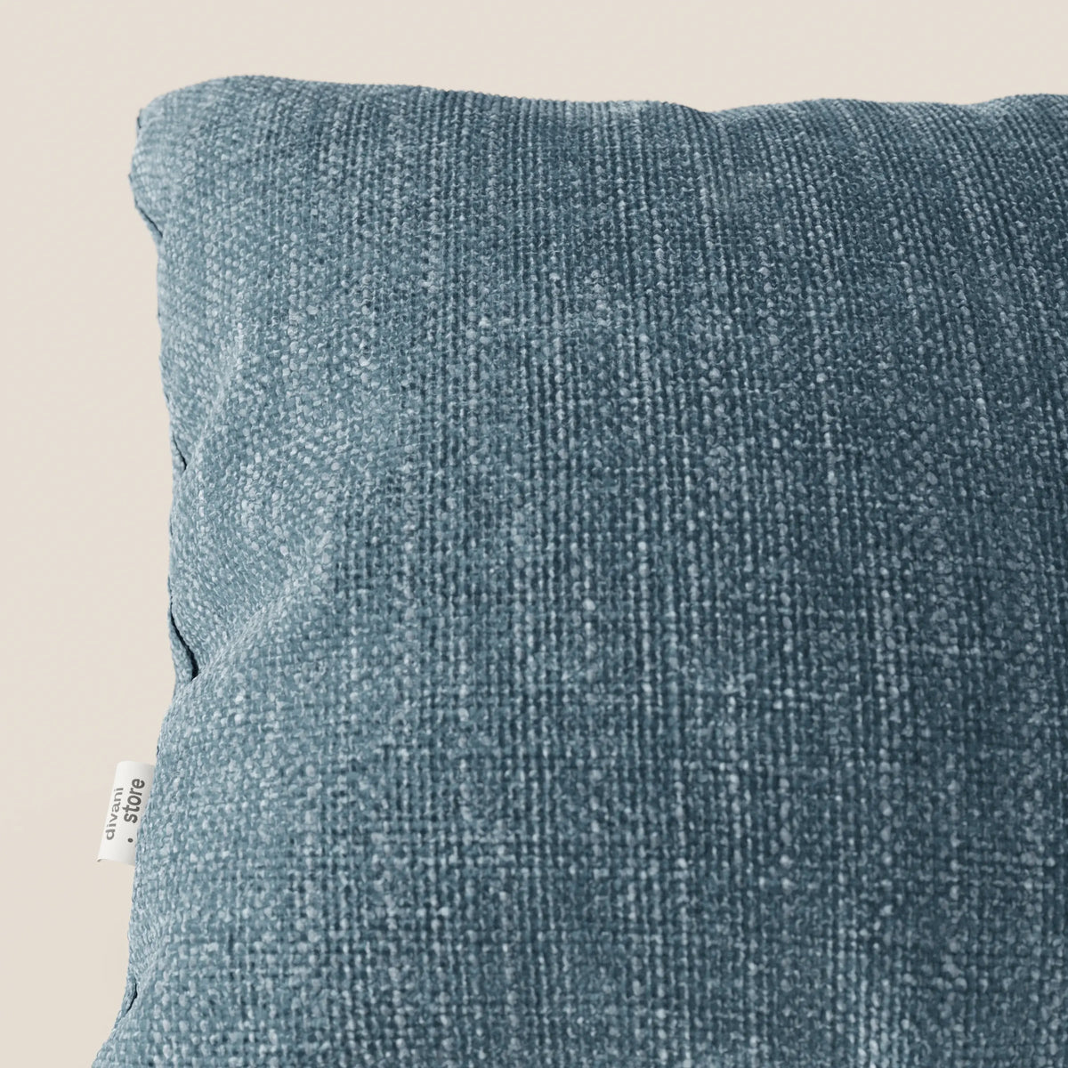Primo piano del cuscino Lyon 45x45 cm in T06 azzurro di divitt.sbs, realizzato in morbido tessuto intrecciato impermeabile, presentato su uno sfondo beige con una piccola etichetta bianca con testo nero sul lato sinistro.