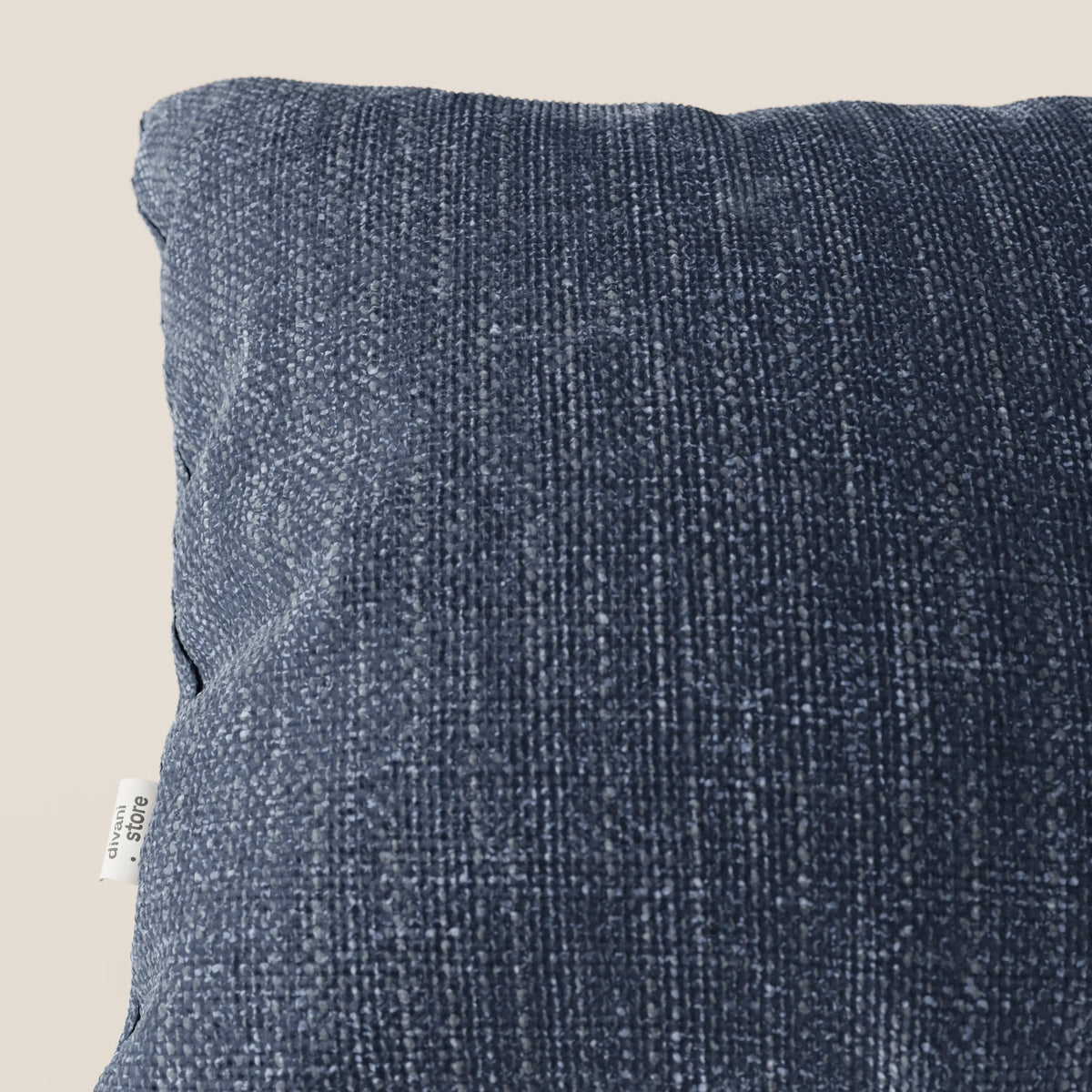 Primo piano del cuscino Lyon di divitt.sbs in T06 blu, realizzato in morbido tessuto impermeabile. Su uno sfondo beige chiaro, sul bordo sinistro compare una piccola etichetta bianca. Il cuscino sembra soffice ed è leggermente inclinato verso la macchina fotografica.