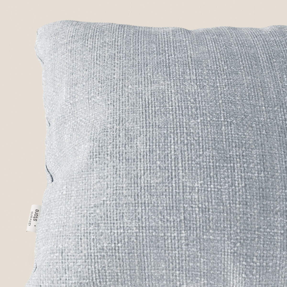 Primo piano del cuscino Lyon 45x45 cm di divitt.sbs in morbido tessuto intrecciato impermeabile T06 ghiaccio, su sfondo beige. Una piccola etichetta di tessuto bianco è visibile sulla cucitura sinistra e ne evidenzia la qualità e la consistenza.