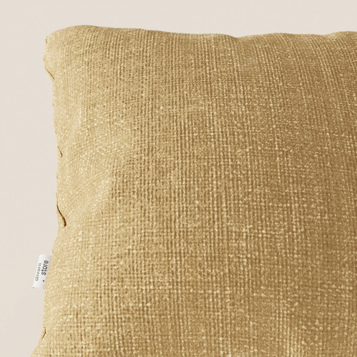 Primo piano del cuscino Lyon 45x45 cm di divitt.sbs in T06 giallo, realizzato in morbido tessuto impermeabile. Su uno sfondo beige chiaro si vede solo una parte del cuscino giallo oro, con una piccola etichetta bianca sul bordo sinistro.