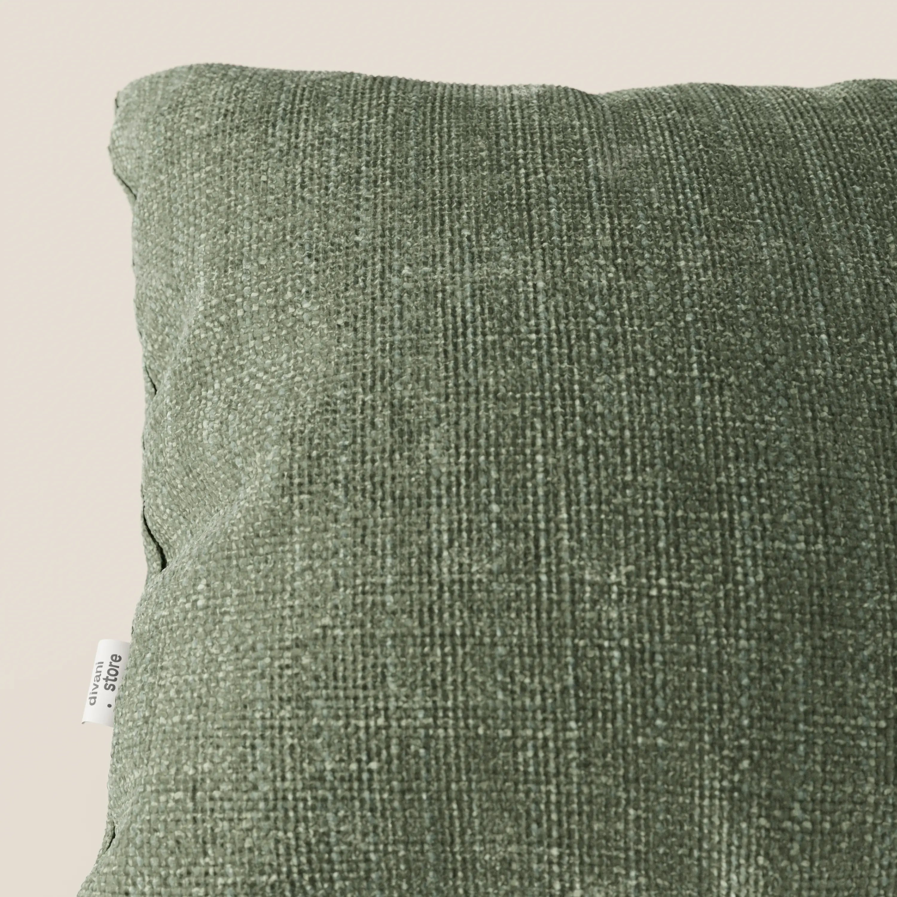 Primo piano del cuscino Lyon 45x45 in tessuto intrecciato impermeabile T06 verde di divitt.sbs, caratterizzato da una morbida trama a quadri e da una piccola etichetta in tessuto bianco sulla sinistra, su uno sfondo beige chiaro.