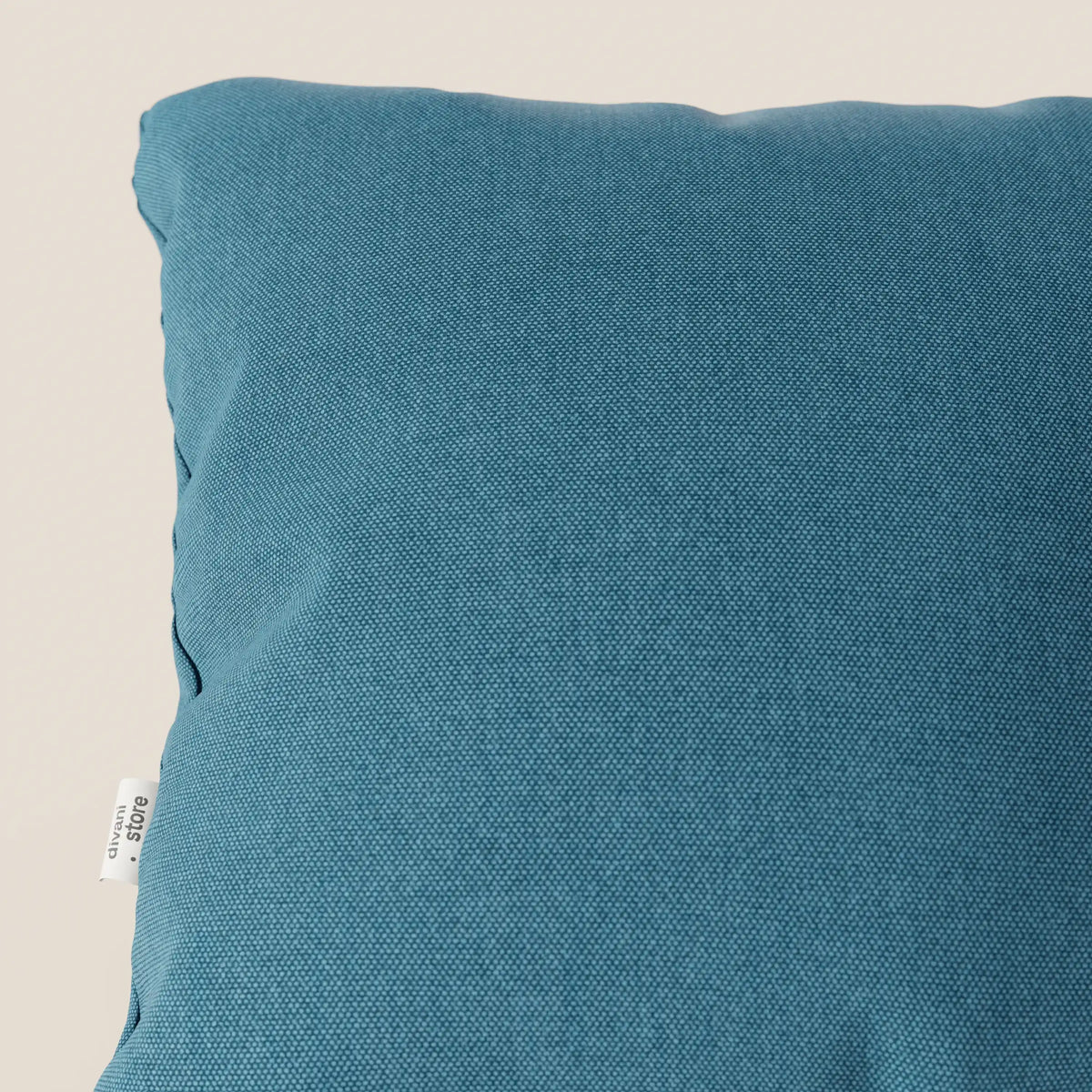 Primo piano del cuscino Honey 45x45 di divitt.sbs in T13 azzurro, caratterizzato da un tessuto simil-cotone impermeabile e da una targhetta laterale bianca. Il fondo beige chiaro ne accentua il design elegante.
