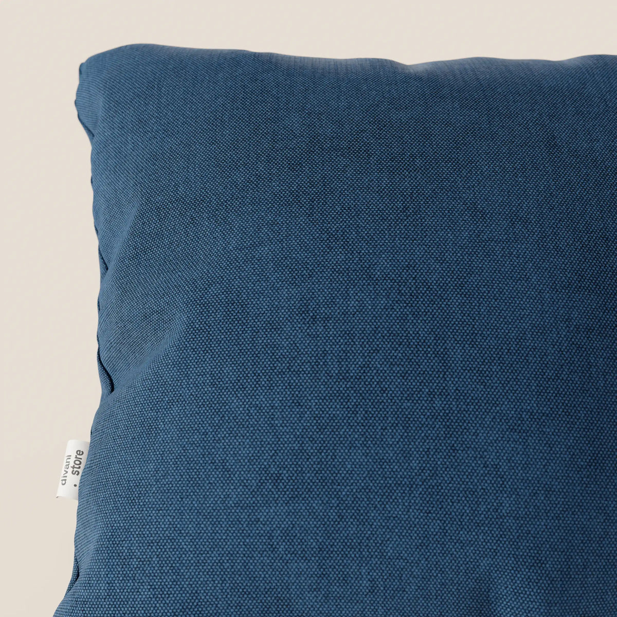 Primo piano del cuscino Honey 45x45 di divitt.sbs, presentato in tessuto blu T13 impermeabile simil-cotone con superficie strutturata e piccola etichetta laterale bianca, pratico ed elegante su uno sfondo beige chiaro.