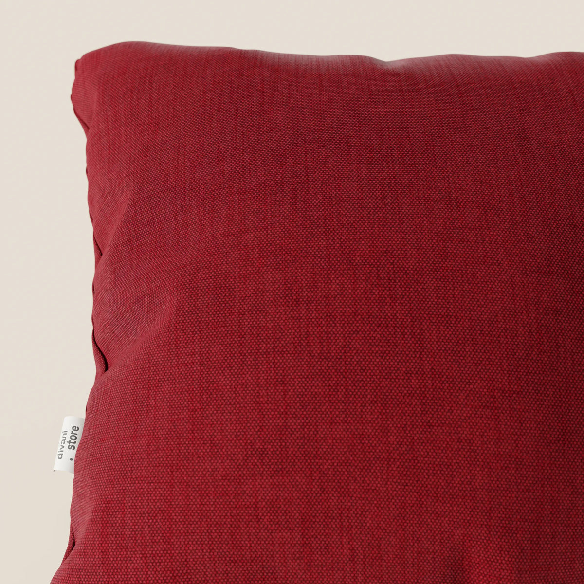 Primo piano del cuscino divitt.sbs Honey 45x45 in T13 rosso tessuto simil cotone impermeabile, che presenta un'etichetta bianca e un sottile motivo a fodera effetto cotone su uno sfondo beige chiaro. È visibile solo una parte del cuscino.