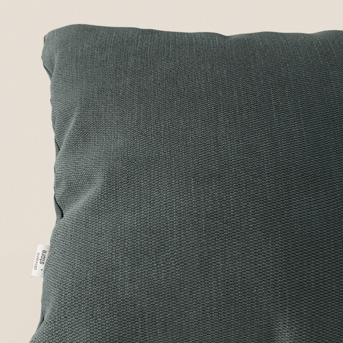 Primo piano di un cuscino Miami 45x45 cm T14 verde di divitt.sbs, in morbido tessuto impermeabile con rivestimento soft-touch, trama intrecciata ed etichetta bianca a vista su sfondo beige chiaro.
