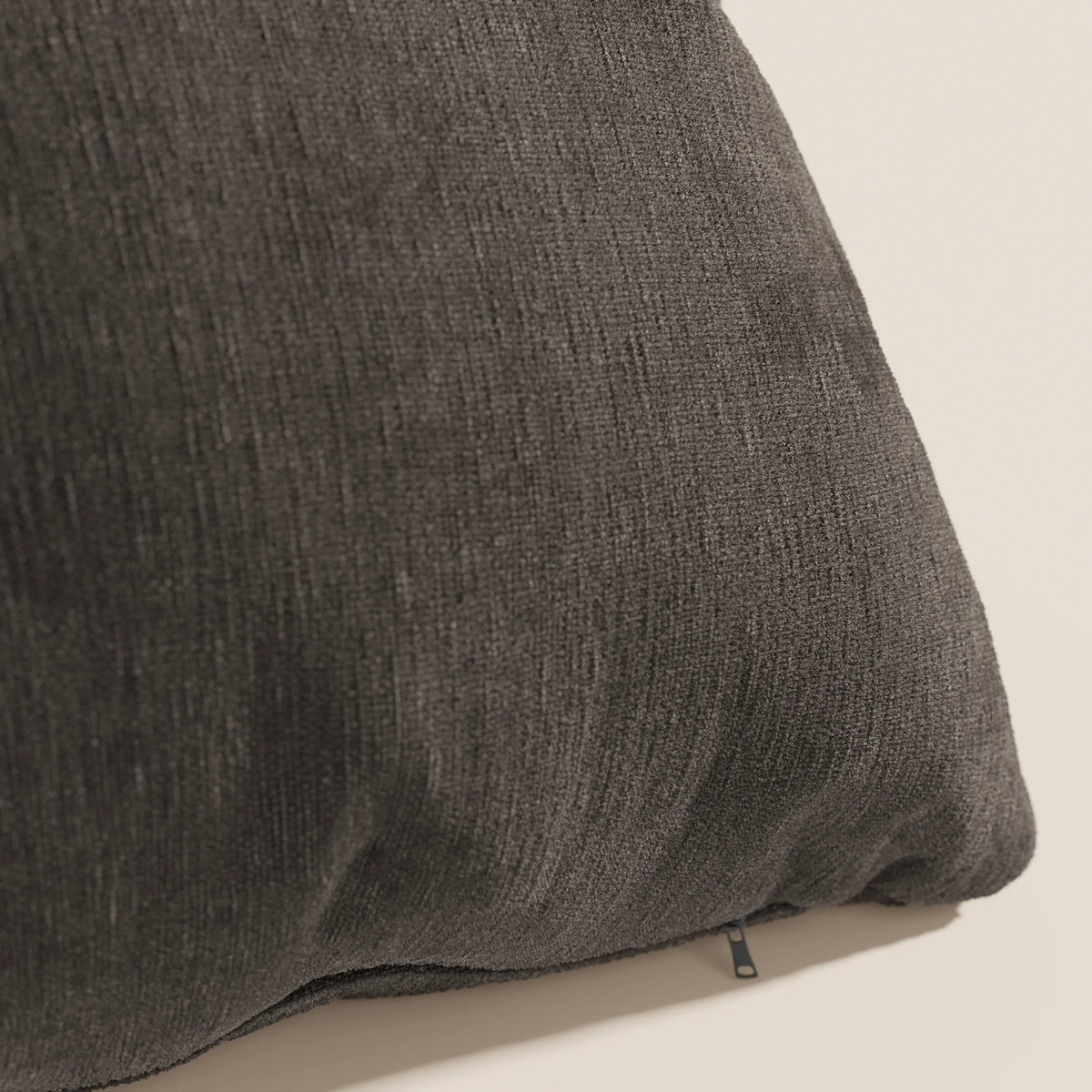 Un primo piano del cuscino Bolero di divitt.sbs in grigio scuro mostra il suo tessuto morbido, intrecciato e smacchiabile T31. Il cuscino è posizionato ad angolo su uno sfondo neutro, con una piccola cerniera nera visibile vicino al bordo inferiore.