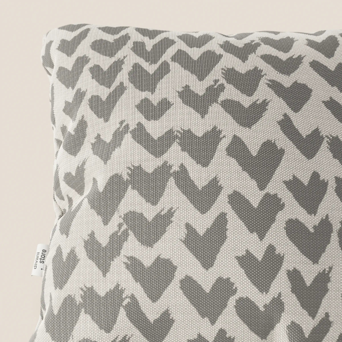 Primo piano dei cuscini geometrici Arrow di divitt.sbs, disponibili in vari colori, realizzati in tessuto idrorepellente grigio. Sul bordo sinistro del cuscino compare una piccola etichetta in tessuto bianco con testo.