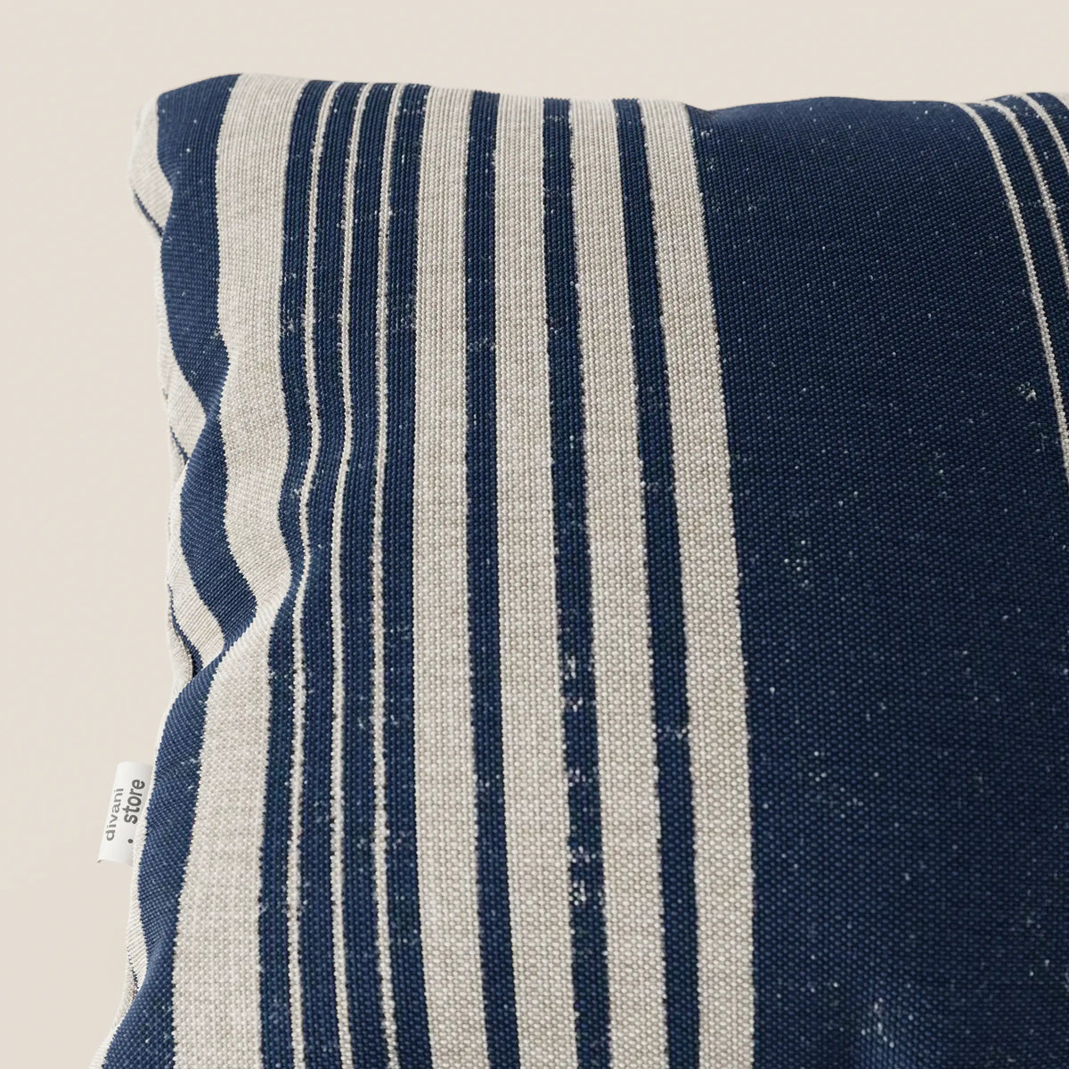 Primo piano del cuscino a fantasia rigata 45x45 di divitt.sbs Line in tessuto impermeabile, blu navy a righe verticali beige. Un cartellino bianco indica "100% cotone". Lo sfondo chiaro evidenzia il motivo decorativo e la trama.