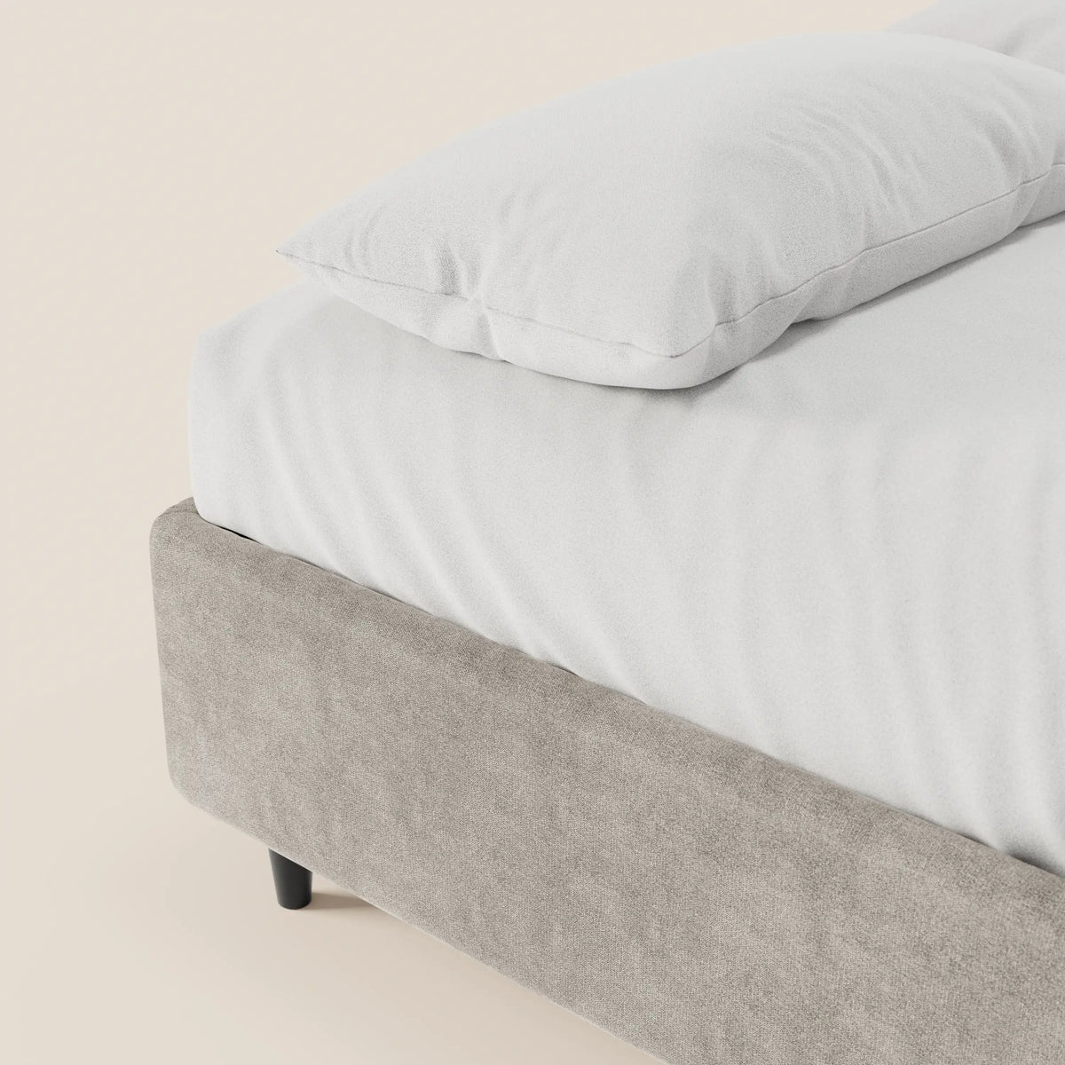 Un primo piano del letto sommier imbottito ERON di Letti.store con un cuscino bianco e un lenzuolo con angoli. La struttura in tessuto Oeko-Tex impermeabile grigio chiaro, le gambe nere e il vano contenitore risaltano su uno sfondo minimalista color avorio.