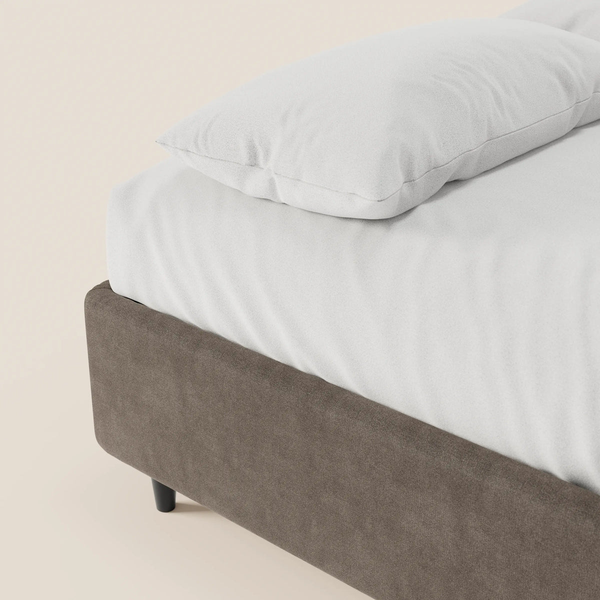 Un primo piano del letto sommier imbottito ERON di Letti.store, con lenzuolo e cuscino bianchi su una struttura rivestita in tessuto marrone simile alla pelle scamosciata, in tessuto impermeabile certificato Oeko-Tex, con gambe corte nere e senza testiera, su uno sfondo color crema chiaro.