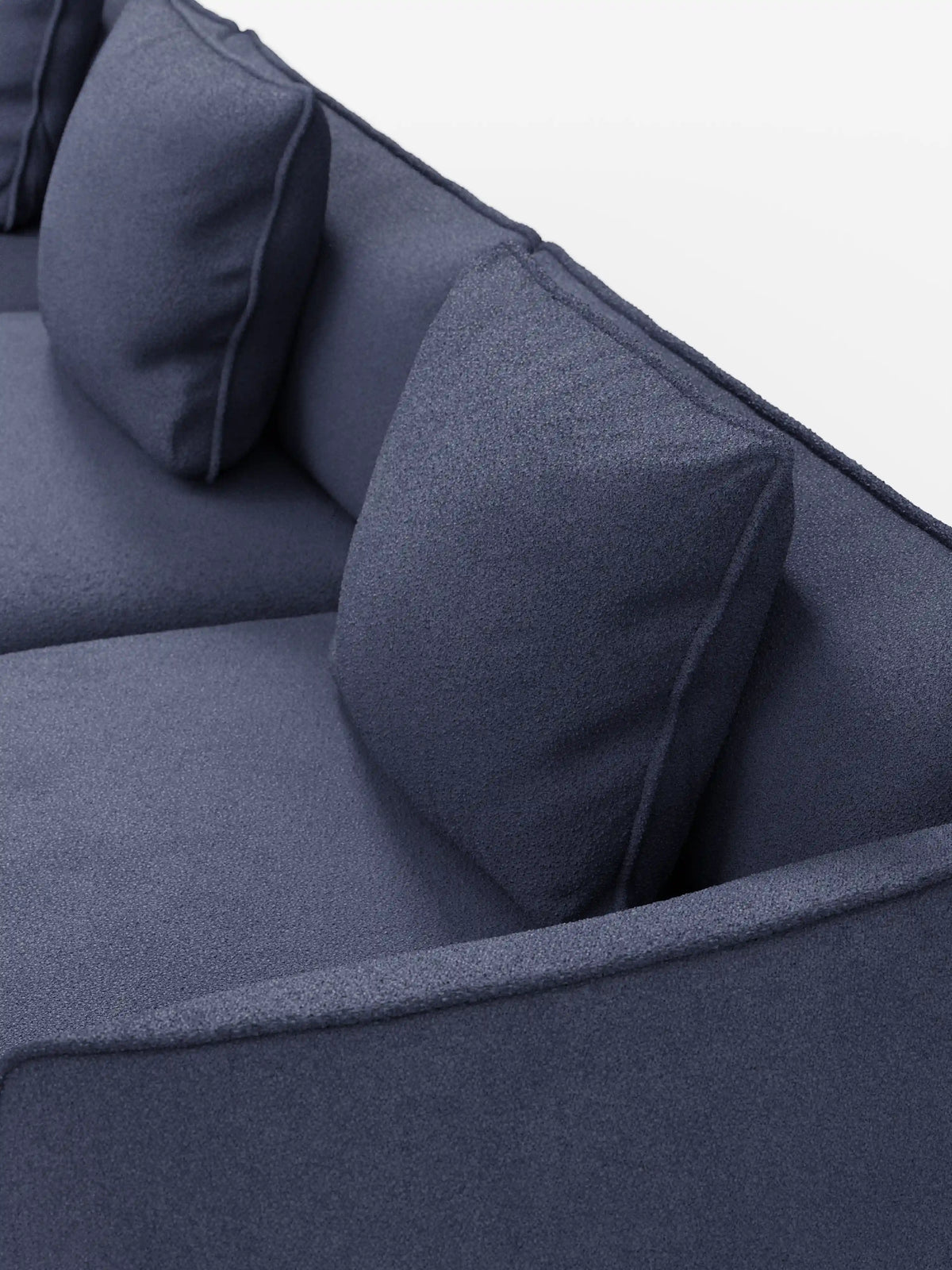 Un primo piano del divano modulare a 3 posti “Nimbus” di divitt.sbs, realizzato in tessuto effetto cotone blu scuro, testurizzato e resistente alle macchie, con due cuscini quadrati per lo schienale, su uno sfondo bianco a tinta unita.