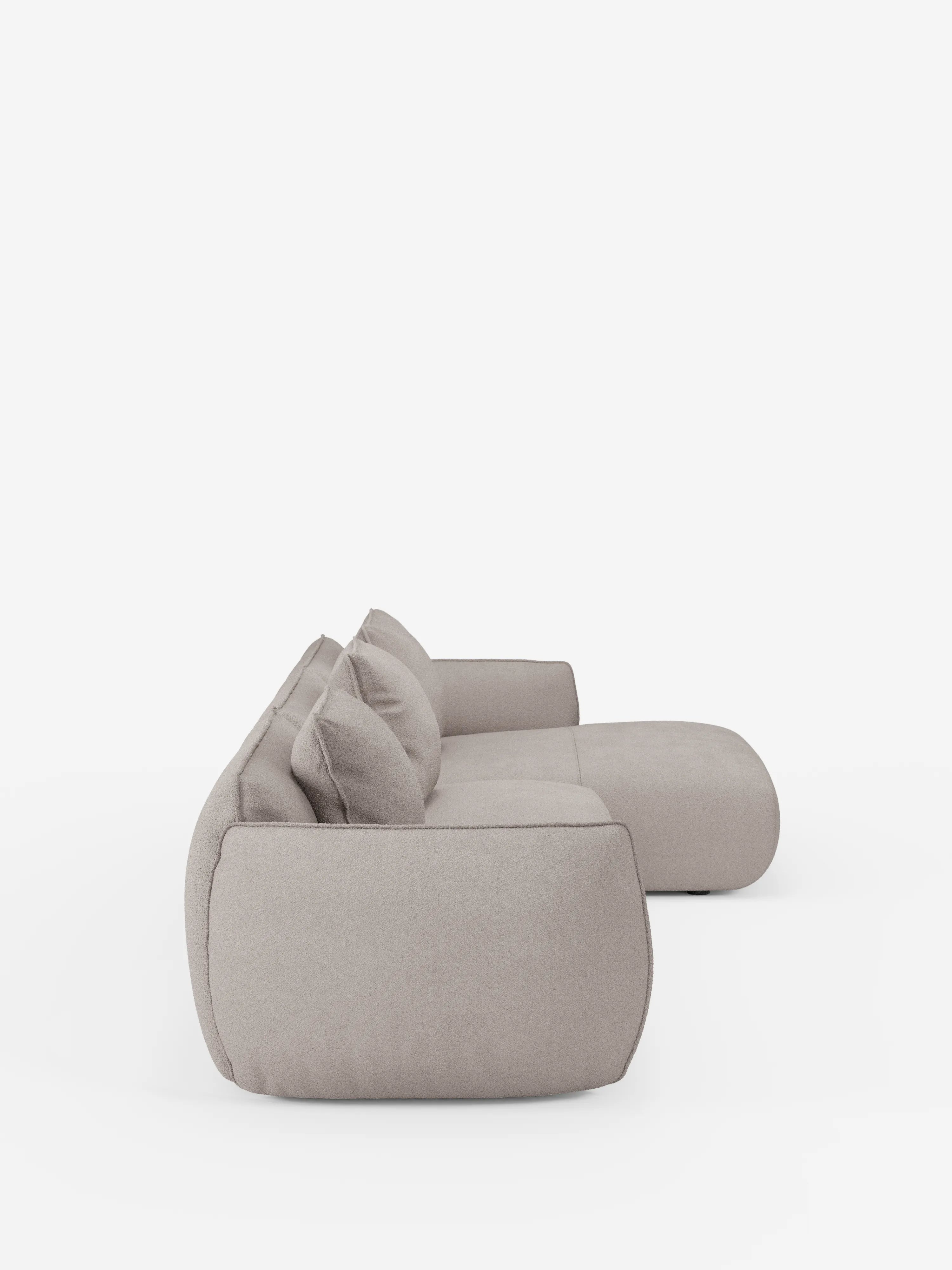 Il divano angolare con penisola componibile Nimbus in morbido tessuto effetto cotone smacchiabile T33 di divitt.sbs è un moderno divano componibile color beige chiaro, ripreso in una vista laterale, caratterizzato da cuscini spessi e arrotondati e da due cuscini dello schienale abbinati. Sullo sfondo bianco a tinta unita, la sua texture morbida e imbottita e il suo design contemporaneo e delicato risaltano ancora di più.