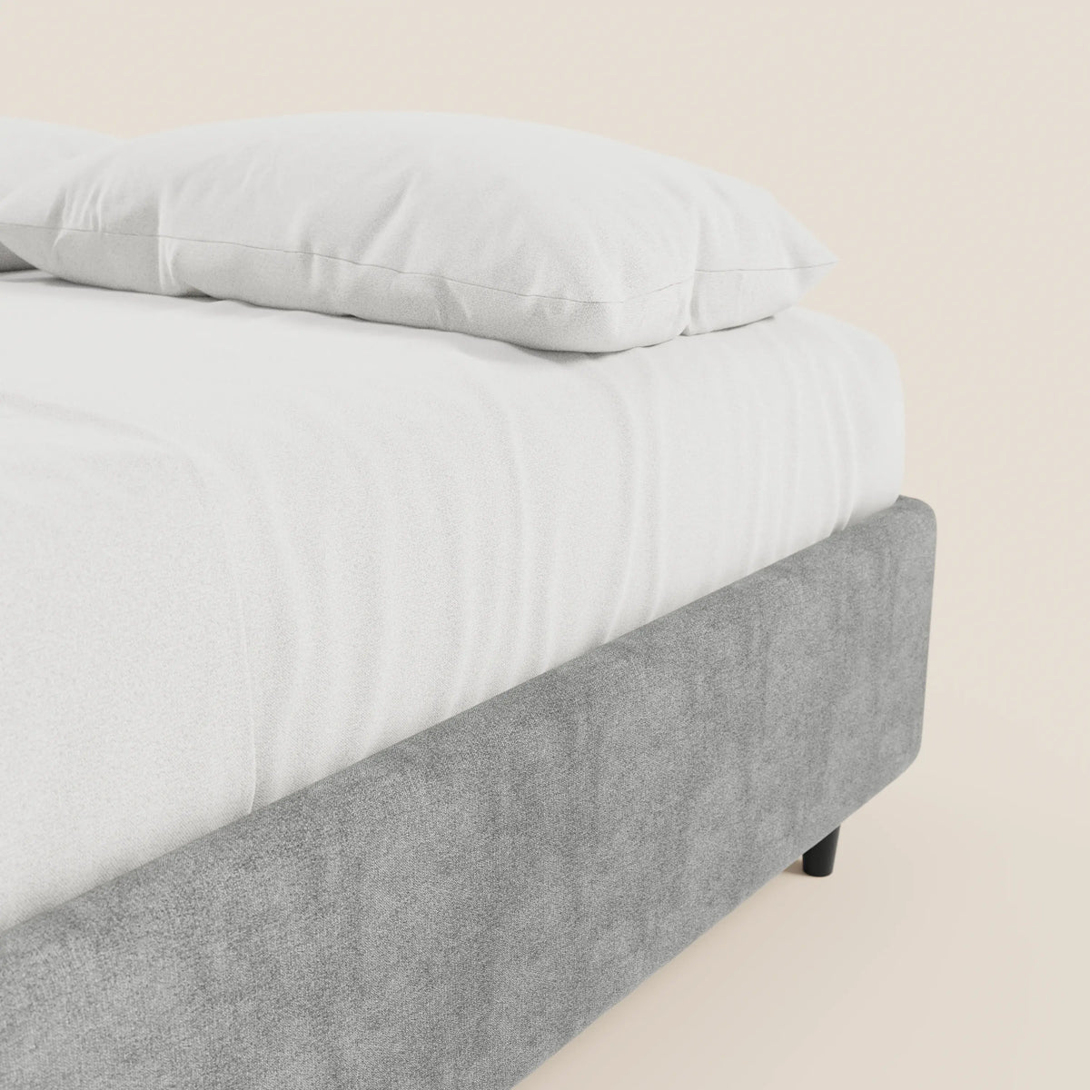 Un primo piano del letto sommier imbottito ERON con contenitore di Letti.store, con cuscino bianco e lenzuolo con angoli. La base in tessuto impermeabile certificato Oeko-Tex, di colore grigio chiaro, e le gambe corte nere risaltano sullo sfondo beige chiaro a tinta unita.