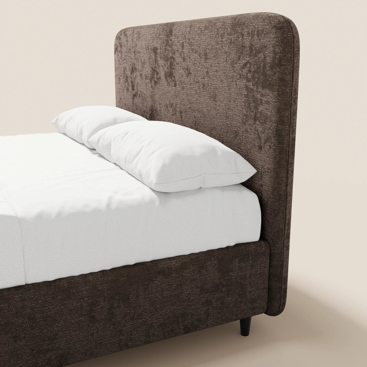 Un primo piano del letto imbottito KAELOS minimal di Letti.store ne mette in risalto la testiera alta e arrotondata in ciniglia e le gambe alte. Rivestito con biancheria da letto di un bianco brillante e due morbidi cuscini, solo una parte di questo letto contenitore è visibile sullo sfondo beige chiaro.