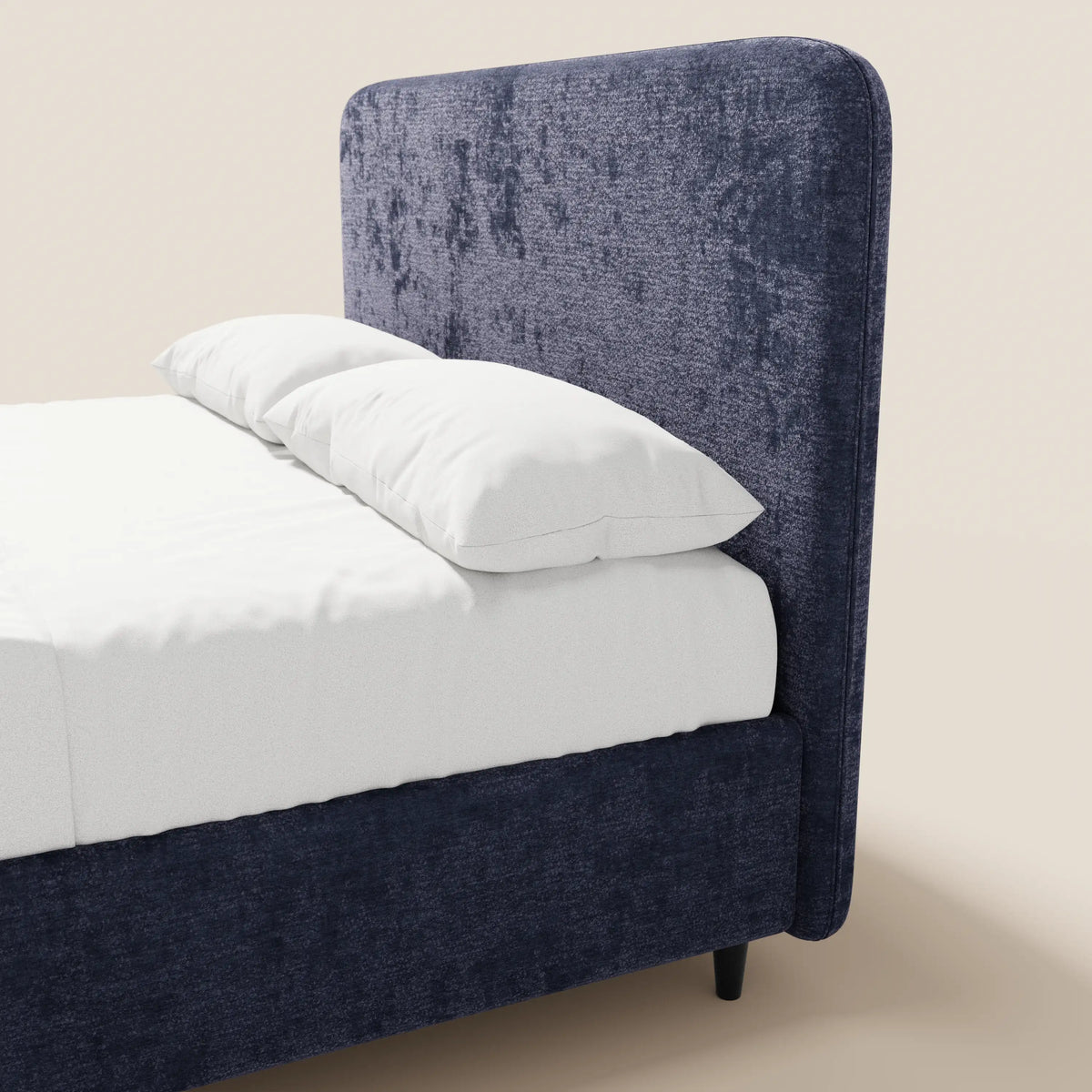 Il letto imbottito KAELOS minimal di Letti.store presenta una testiera arrotondata in ciniglia, gambe alte e un vano contenitore integrato. Nella foto è raffigurato in blu navy con lenzuola e cuscini di un bianco candido, su uno sfondo beige chiaro.