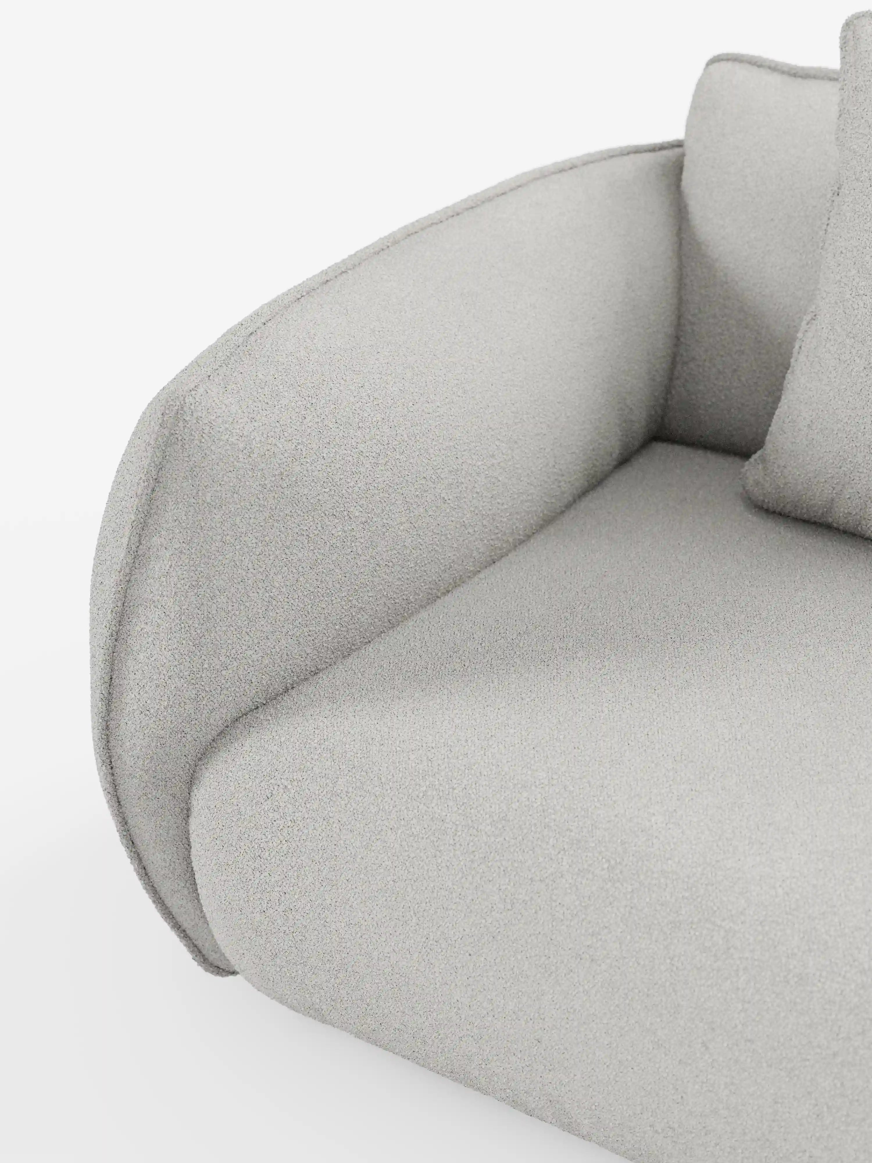Primo piano del divano angolare con penisola componibile Nimbus di divitt.sbs, rivestito in tessuto T33 grigio chiaro, morbido, dall’effetto cotone e resistente alle macchie. L'immagine mostra il bracciolo ricurvo di questo divano moderno, il cuscino spesso e un set di cuscini coordinati su uno sfondo bianco, mettendo in risalto la trama morbida e leggermente felpata che garantisce un comfort eccezionale.