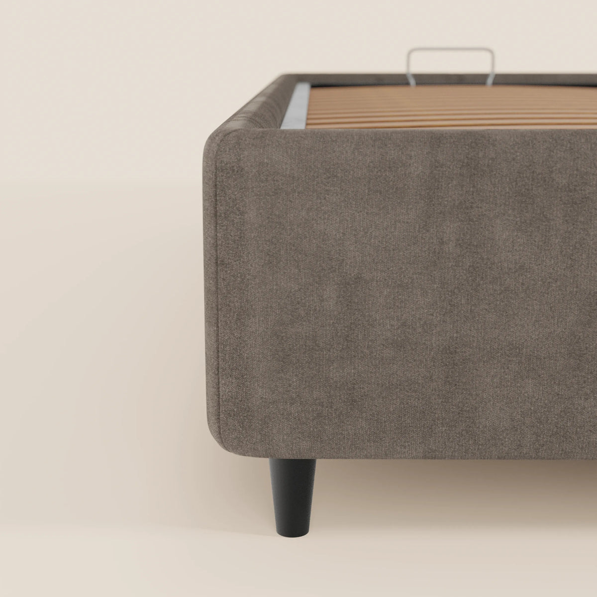 Un primo piano del letto sommier imbottito ERON di Letti.store ne mostra il tessuto grigio impermeabile certificato Oeko-Tex, le gambe affusolate nere e le doghe in legno a vista su uno sfondo liscio color beige chiaro, con il letto posizionato sul lato destro dell'immagine.