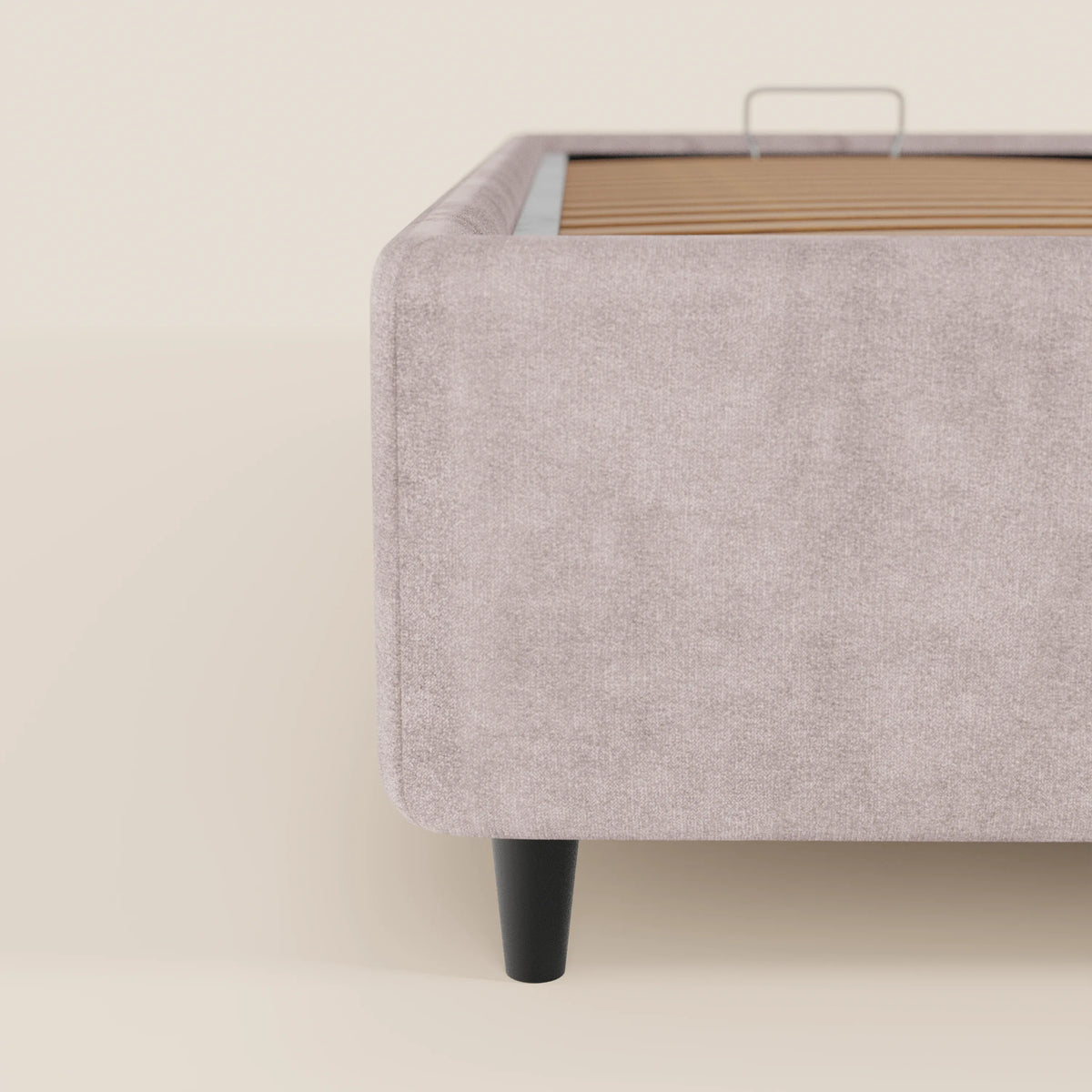 Primo piano della vista laterale del letto sommier imbottito ERON di Letti.store, caratterizzato da un tessuto beige chiaro impermeabile certificato Oeko-Tex, doghe in legno, un’unica gamba nera affusolata e un ampio vano contenitore. L’estetica minimalista risalta sullo sfondo coordinato.