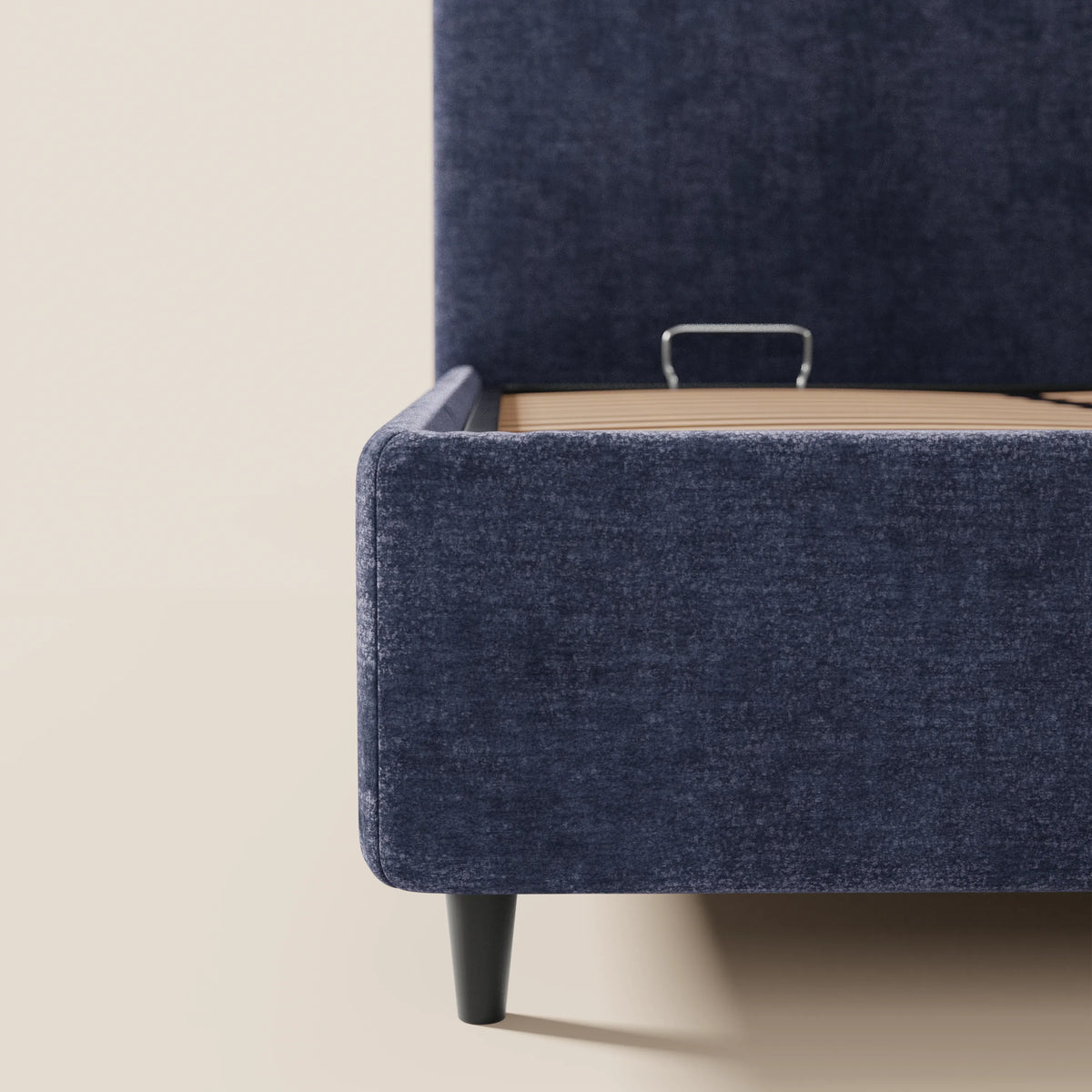 Primo piano del letto KAELOS di Letti.store, un modello minimal imbottito in ciniglia blu scuro, caratterizzato da una testiera arrotondata, gambe alte e vano contenitore. La struttura mette in risalto le doghe in legno e il supporto metallico per il materasso su uno sfondo chiaro e uniforme.