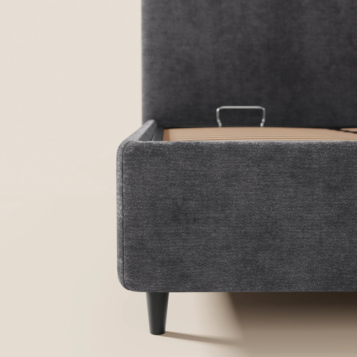 Primo piano del letto imbottito KAELOS minimal di Letti.store, caratterizzato da una testiera alta e arrotondata, gambe alte e rivestimento in ciniglia grigio scuro con vano portaoggetti, ripreso su un pavimento chiaro. All’interno del telaio del letto è visibile una barra metallica di fissaggio del materasso.