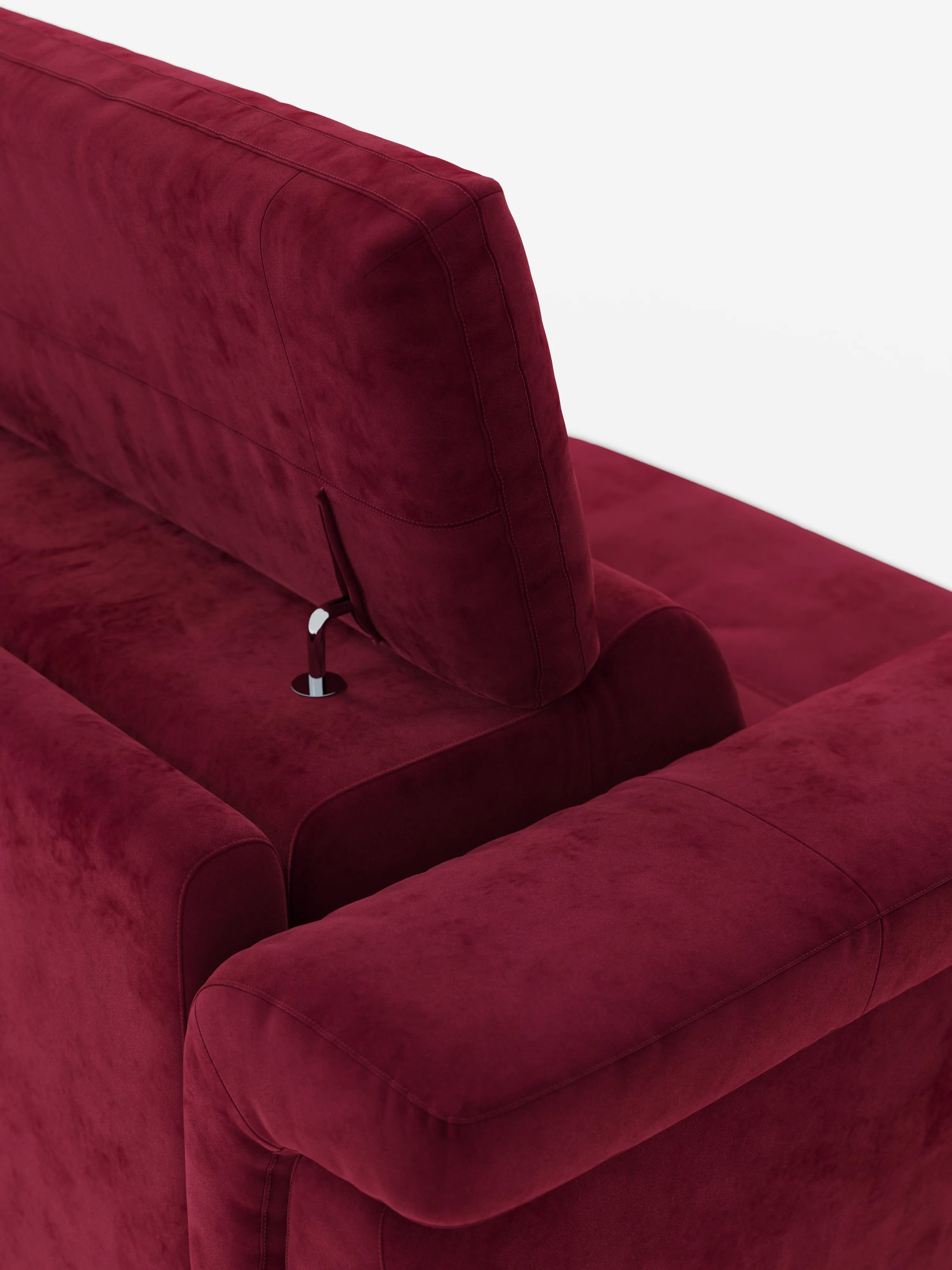 Un primo piano dello schienale e del bracciolo del divano letto di design Capri con piedini alti di divitt.sbs mette in risalto il suo rivestimento in morbido tessuto effetto velluto smacchiabile di un rosso intenso (tessuto effetto velluto T30). Una leva metallica situata vicino al poggiatesta regolabile lascia intuire le sue caratteristiche ergonomiche. Su uno sfondo bianco a tinta unita, le finiture morbide e lussuose del divano e le cuciture pulite incarnano la classica maestria artigianale italiana.