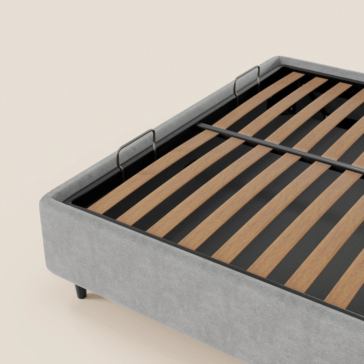 Un primo piano del letto sommier imbottito ERON di Letti.store, caratterizzato da una base in tessuto impermeabile Oeko-Tex grigio chiaro, gambe in metallo nero, doghe in legno di sostegno e due staffe metalliche a forma di U agli angoli. Lo sfondo è di colore beige chiaro a tinta unita.