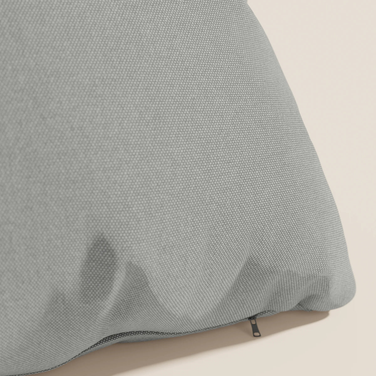 Primo piano del cuscino Honey 45x45 cm di divitt.sbs in impermeabile T13 grigio simil cotone, caratterizzato da una superficie morbida e da una cerniera nera parzialmente visibile lungo un bordo, su sfondo beige.