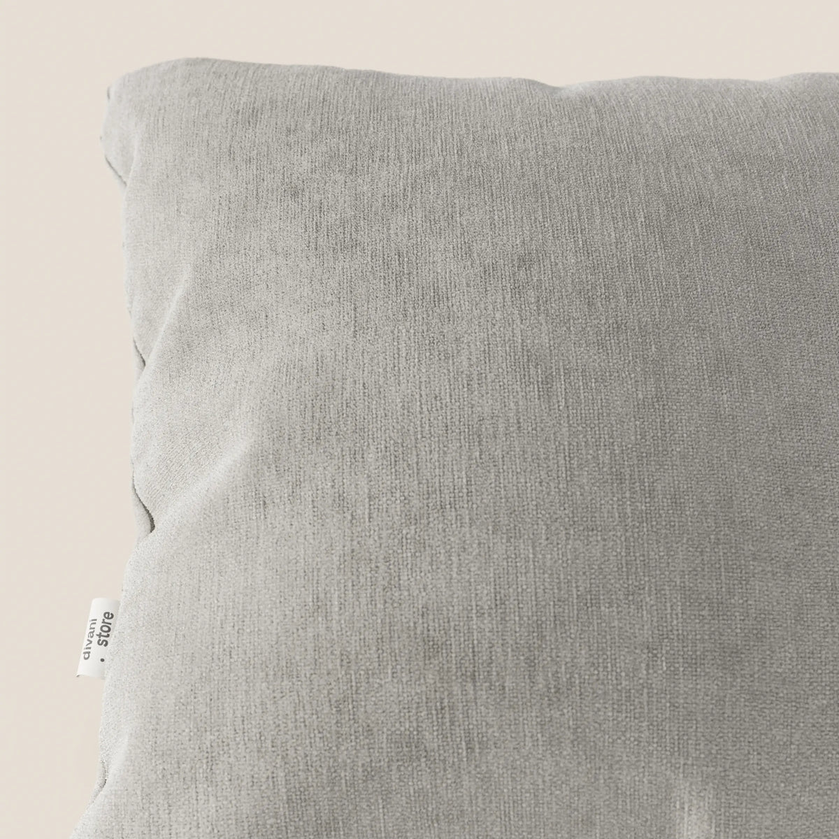 Primo piano del cuscino Bolero di divitt.sbs in grigio chiaro, realizzato in morbido tessuto T31 antimacchia, impermeabile e non restringibile, su sfondo beige con una piccola etichetta bianca sulla cucitura in basso a sinistra.