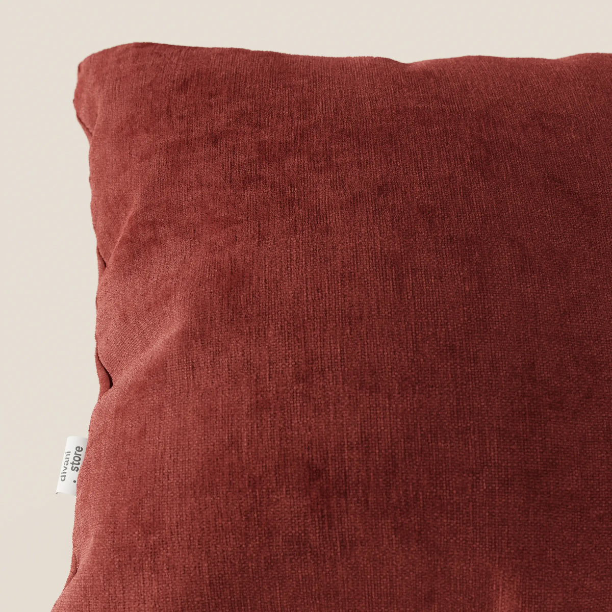 Primo piano del cuscino Bolero di divitt.sbs in tessuto T31 marrone intenso, smacchiabile, impermeabile e irrestringibile, quadrato e strutturato con una piccola etichetta bianca, su sfondo beige chiaro. È visibile solo una parte del cuscino.