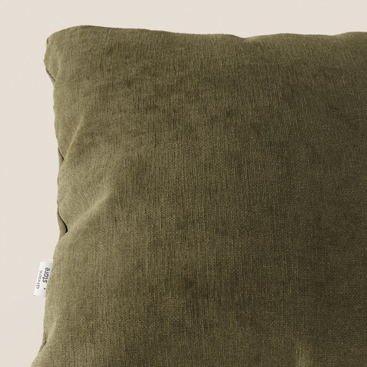 Primo piano di un cuscino Bolero quadrato verde oliva di divitt.sbs in morbido tessuto T31, resistente alle macchie, impermeabile e non restringente, con una sottile trama a coste; piccola etichetta laterale bianca, su sfondo beige chiaro.