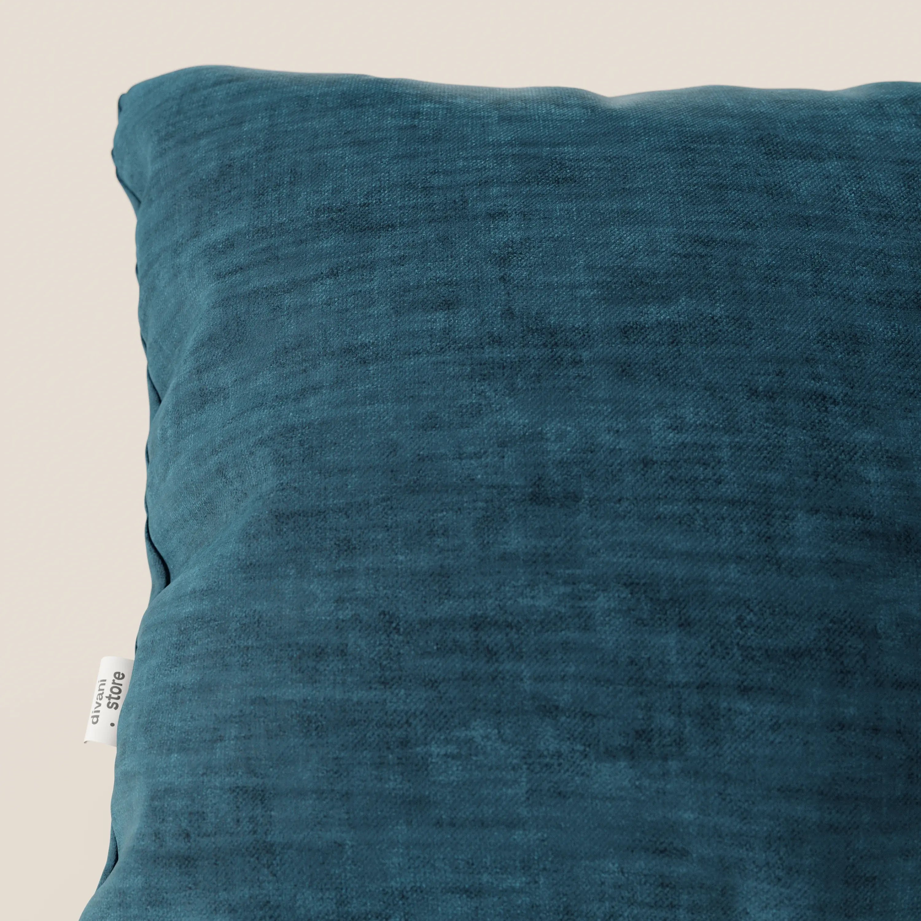 Primo piano del cuscino Sofia di divitt.sbs, presentato in velluto blu acqua quadrato (morbido velluto nuvolato smacchiabile e impermeabile Easy Clean T35), posizionato in diagonale con una piccola etichetta bianca, su uno sfondo beige chiaro.