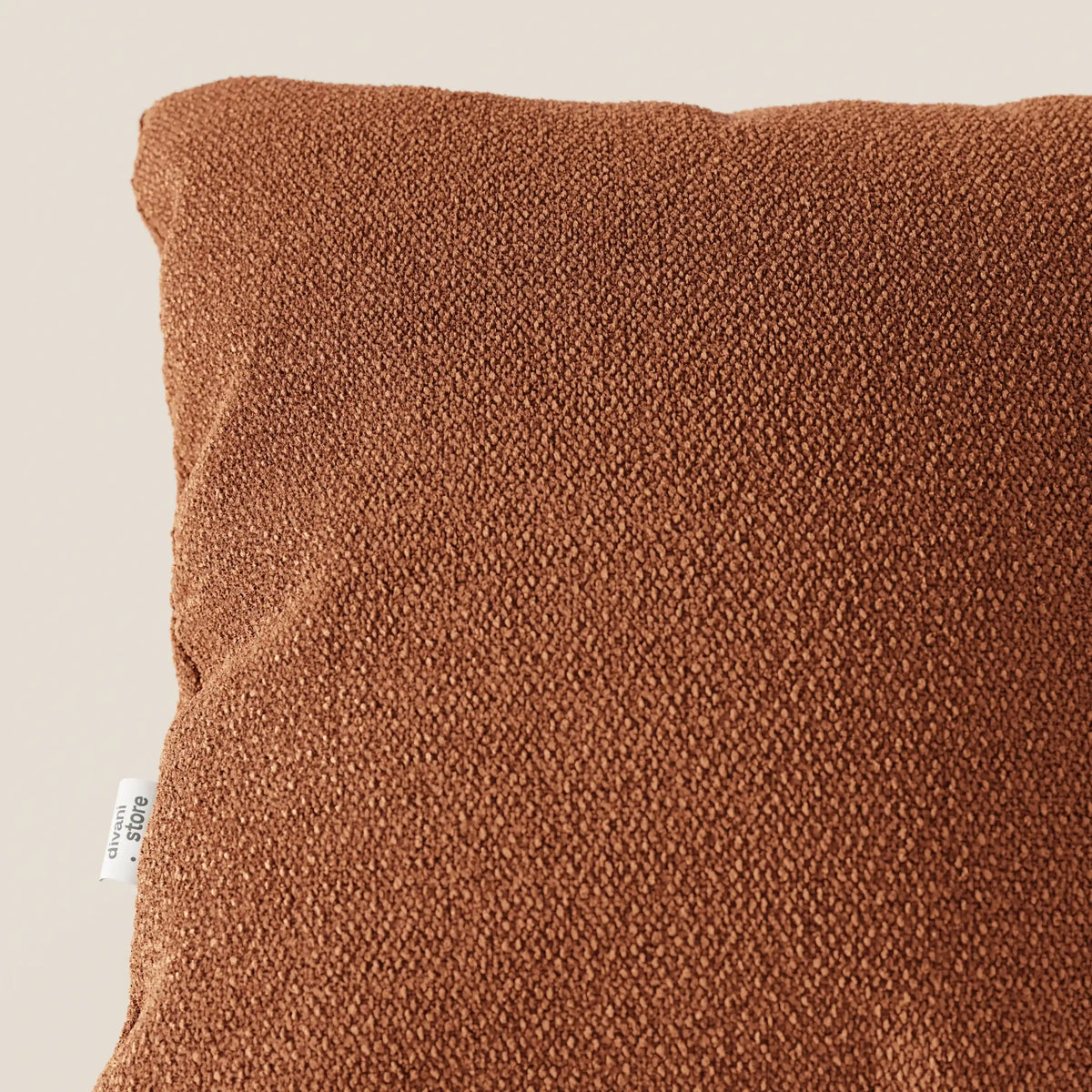 Primo piano dell'angolo superiore sinistro di un cuscino Lambada marrone di divitt.sbs, realizzato in tessuto smacchiabile e impermeabile tipo cotone naturale. Una piccola etichetta bianca con testo nero è visibile sul bordo sinistro del cuscino su uno sfondo neutro.