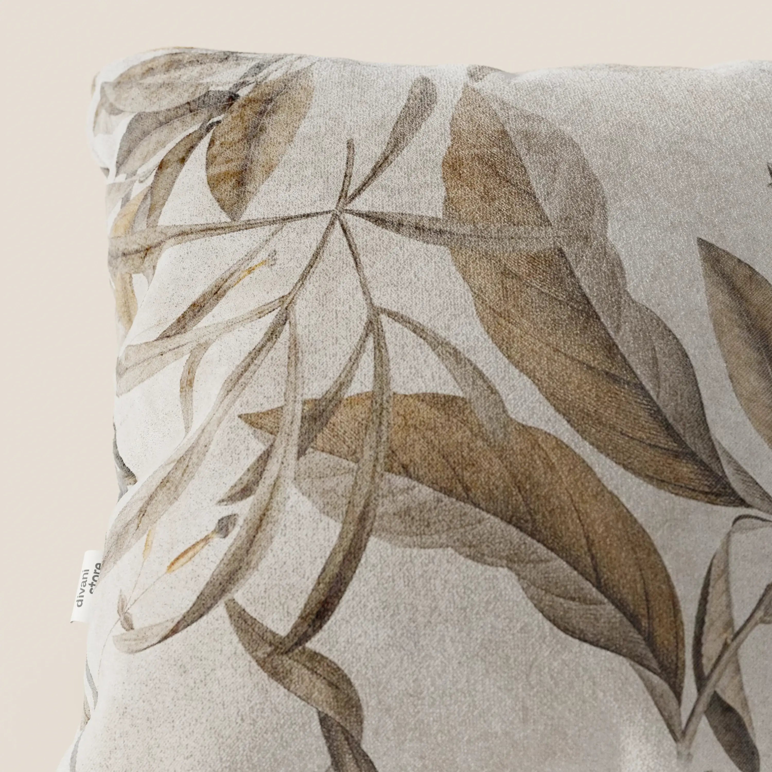 Primo piano del cuscino Lemon elegante a fantasia in velluto impermeabile beige di divitt.sbs, caratterizzato da grandi foglie botaniche marroni e abbronzate su sfondo beige chiaro, texture vellutata e una piccola etichetta bianca sulla cucitura sinistra.