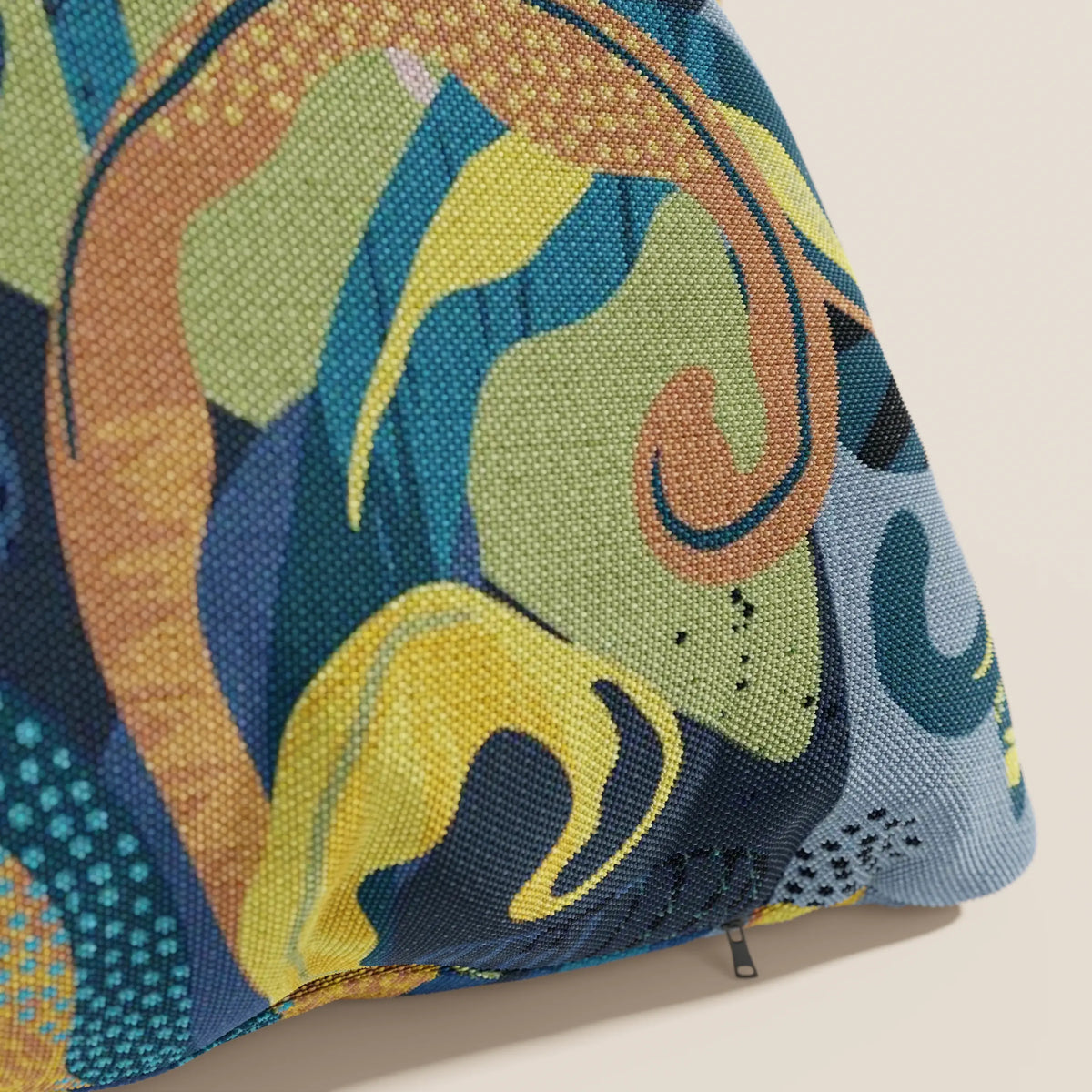 Un primo piano di Joy collezione cuscini a fantasie multicolor di divitt.sbs in fresco cotone impermeabile blu mostra un tessuto strutturato con motivi astratti blu, verdi, gialli, marroni e verde acqua. In basso a destra compare una piccola cerniera.