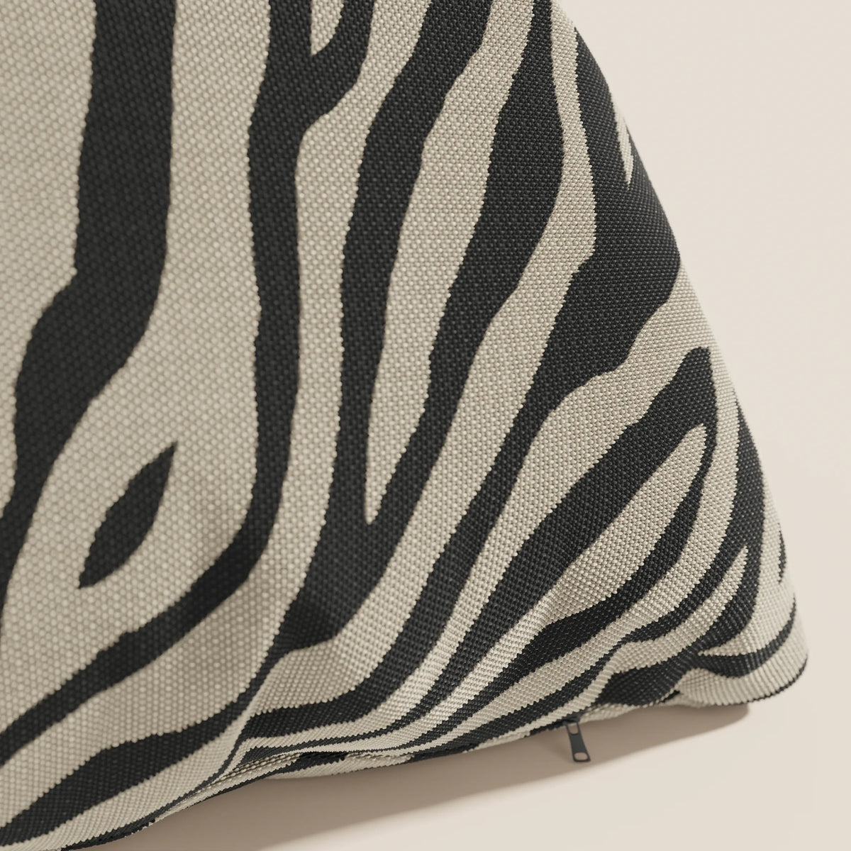 Primo piano di un cuscino divitt.sbs Stripe geometrico in tessuto idrorepellente color tortora a righe zebrate nere e beige, che mostra la trama intrecciata e una piccola chiusura lampo nera su uno sfondo chiaro.