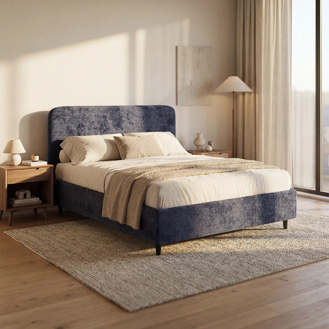 Una camera da letto moderna è arredata con il letto KAELOS minimal imbottito di Letti.store in blu navy, con biancheria da letto bianca e un copriletto beige. Due comodini in legno con lampade fiancheggiano il letto, mentre la luce si riversa su un tappeto intrecciato posto sotto un quadro astratto appeso alla parete.