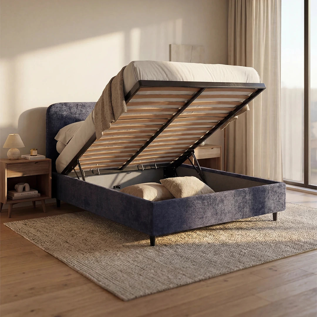 Una camera da letto moderna è arredata con il letto KAELOS minimal imbottito di Letti.store, dotato di vano contenitore, piedini alti e testiera arrotondata in ciniglia. Pavimenti in legno, un tappeto beige, comodini in legno con lampade e ampie finestre inondate di luce completano l'ambiente.
