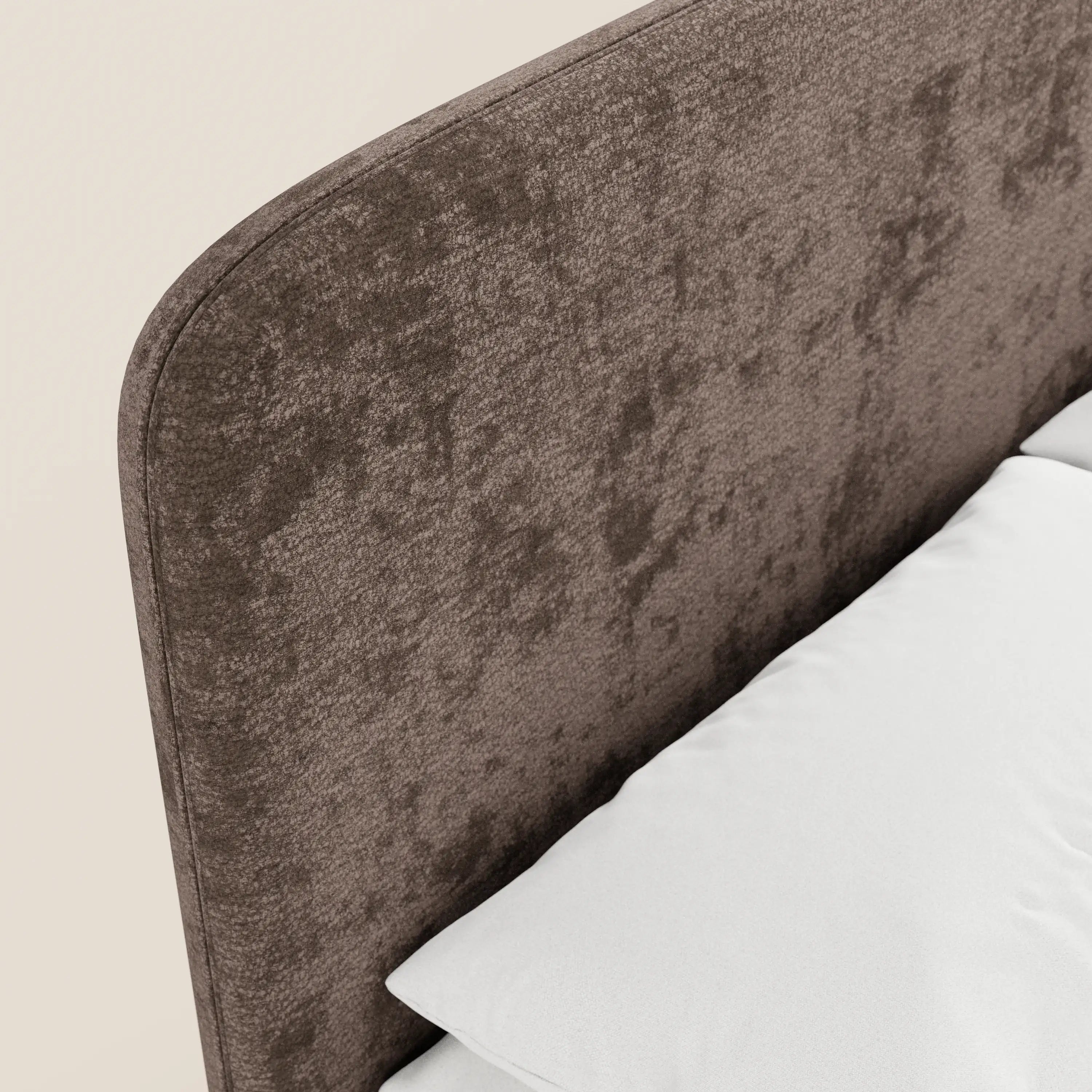 Un primo piano del letto imbottito minimal KAELOS di Letti.store mette in risalto la testiera arrotondata in ciniglia, i piedini alti e il vano portaoggetti, accanto alla biancheria da letto e al cuscino bianchi, su uno sfondo beige a tinta unita.