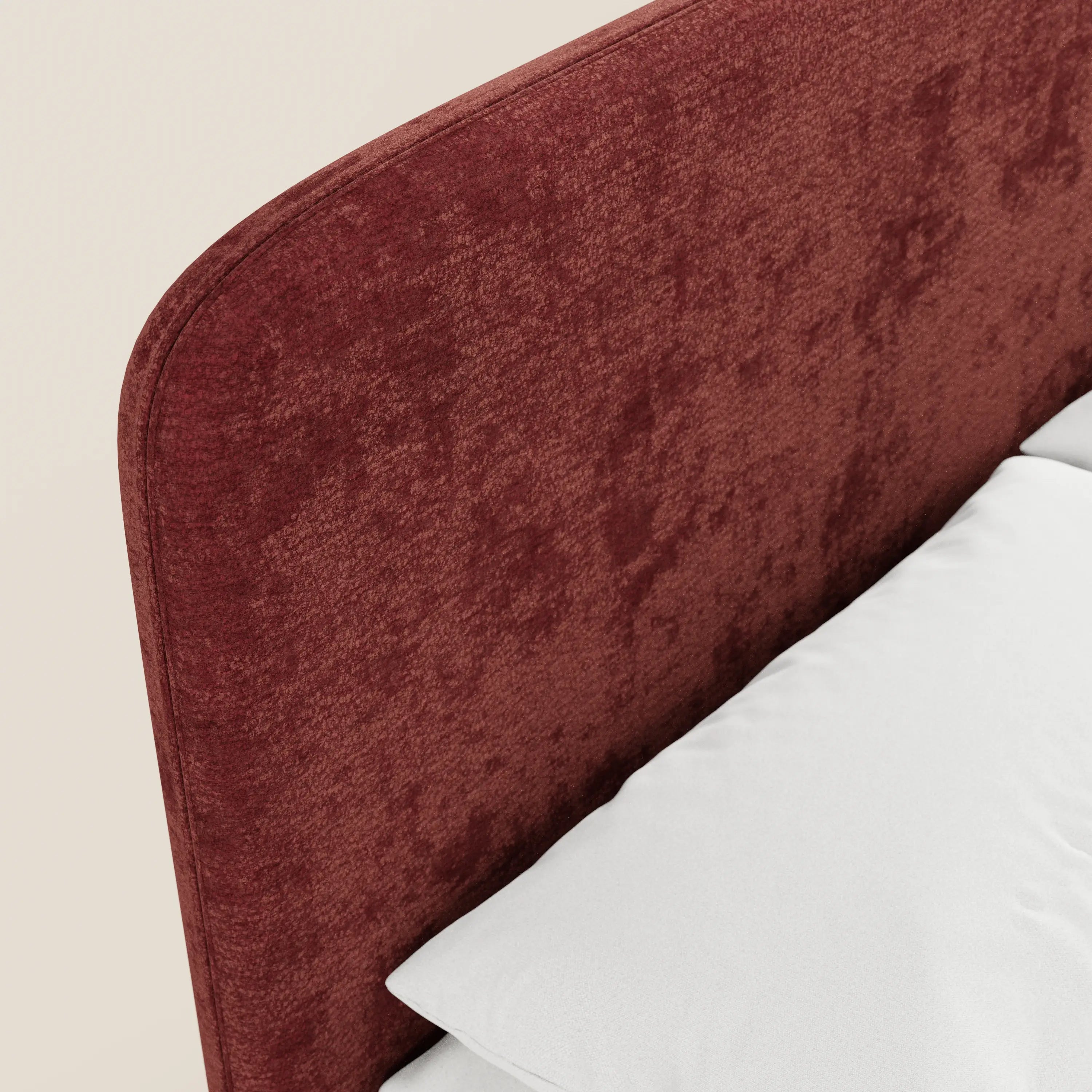 Primo piano del letto imbottito KAELOS Letto Minimal di Letti.store, caratterizzato da una testiera rivestita in ciniglia rosso scuro, gambe alte, design arrotondato, cuscino e materasso bianchi, su uno sfondo beige chiaro.