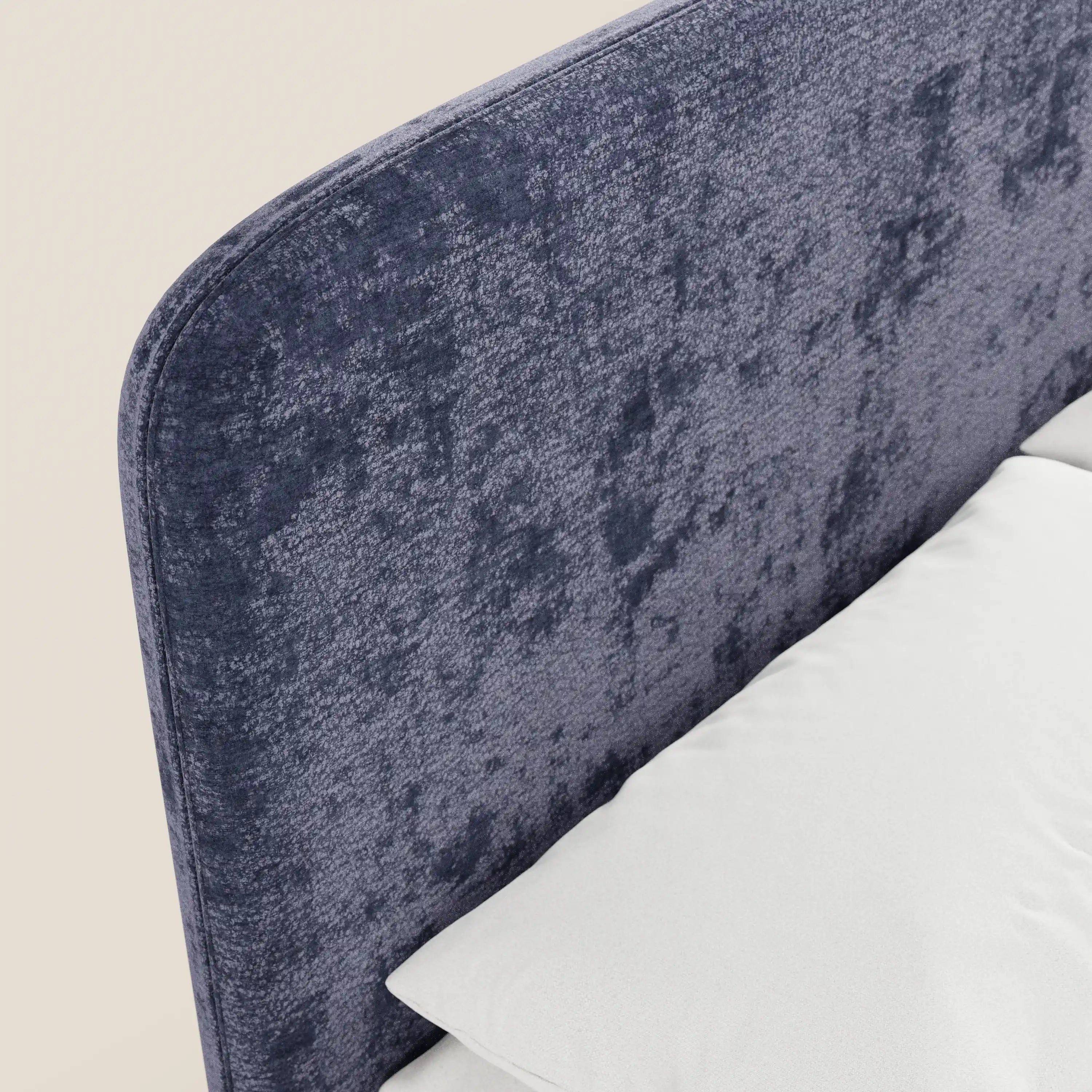 Un primo piano del letto imbottito KAELOS minimal di Letti.store mette in risalto la testiera in ciniglia blu scuro con texture e angoli arrotondati, le gambe alte e la base contenitiva. In basso a destra, su uno sfondo neutro, si intravedono la biancheria da letto bianca e un cuscino.