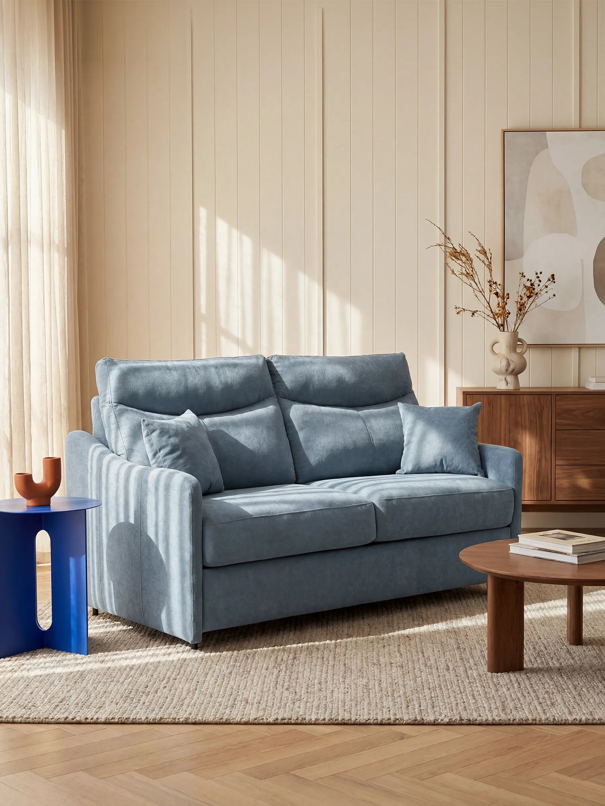 Il divano letto Lodovico a 3 posti con materasso ALTO 20 cm in tessuto smacchiabile certificato Oeko-Tex di divitt.sbs, rivestito in tessuto azzurro, è disposto con due cuscini su un tappeto beige a trama ruvida. La stanza inondata di luce, con pareti rivestite di pannelli color crema, presenta anche un tavolino rotondo in legno, un piccolo tavolino blu su cui è posato un vaso marrone e una credenza in legno decorata con opere d’arte astratta e piante essiccate.