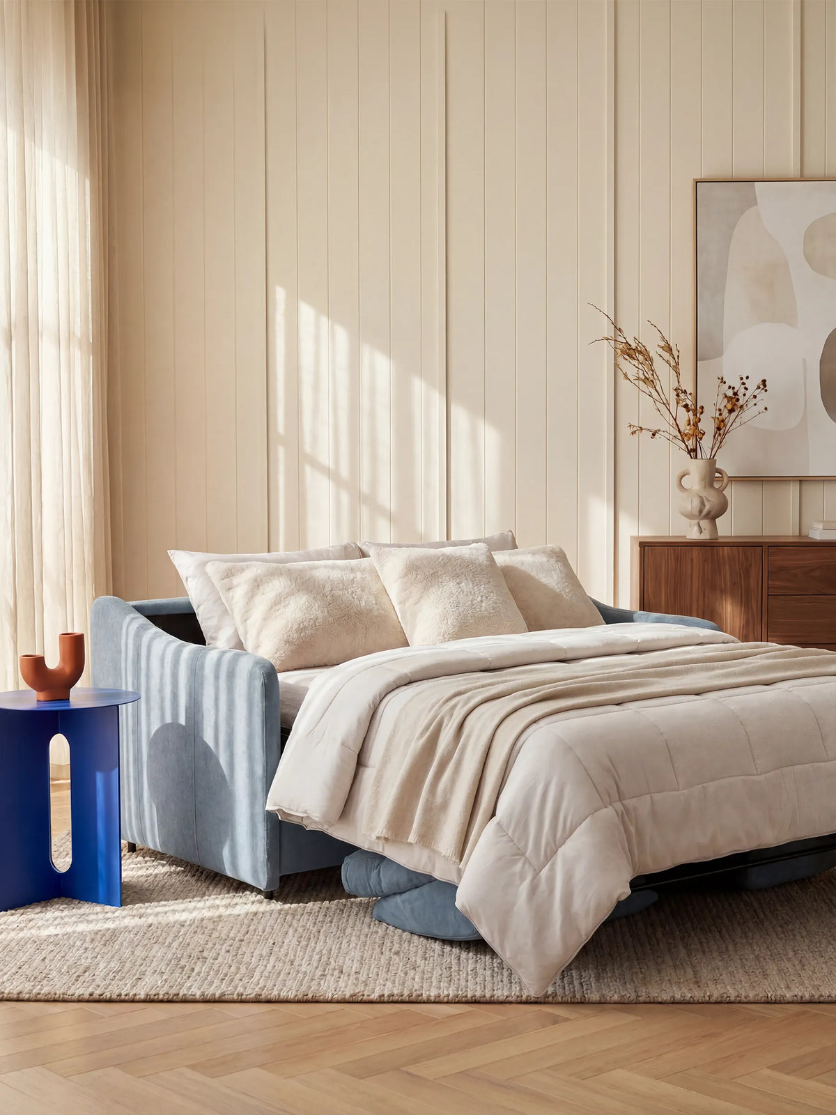 Una camera da letto accogliente è arredata con il divano letto Lodovico a 3 posti con materasso ALTO 20 cm in tessuto smacchiabile certificato Oeko-Tex di divitt.sbs, in azzurro, con biancheria da letto e cuscini beige, posizionato su un tappeto intrecciato. Un tavolino blu con elementi decorativi, una cassettiera in legno, un quadro astratto e un vaso con rami secchi completano l’ambiente moderno e illuminato da luci soffuse.