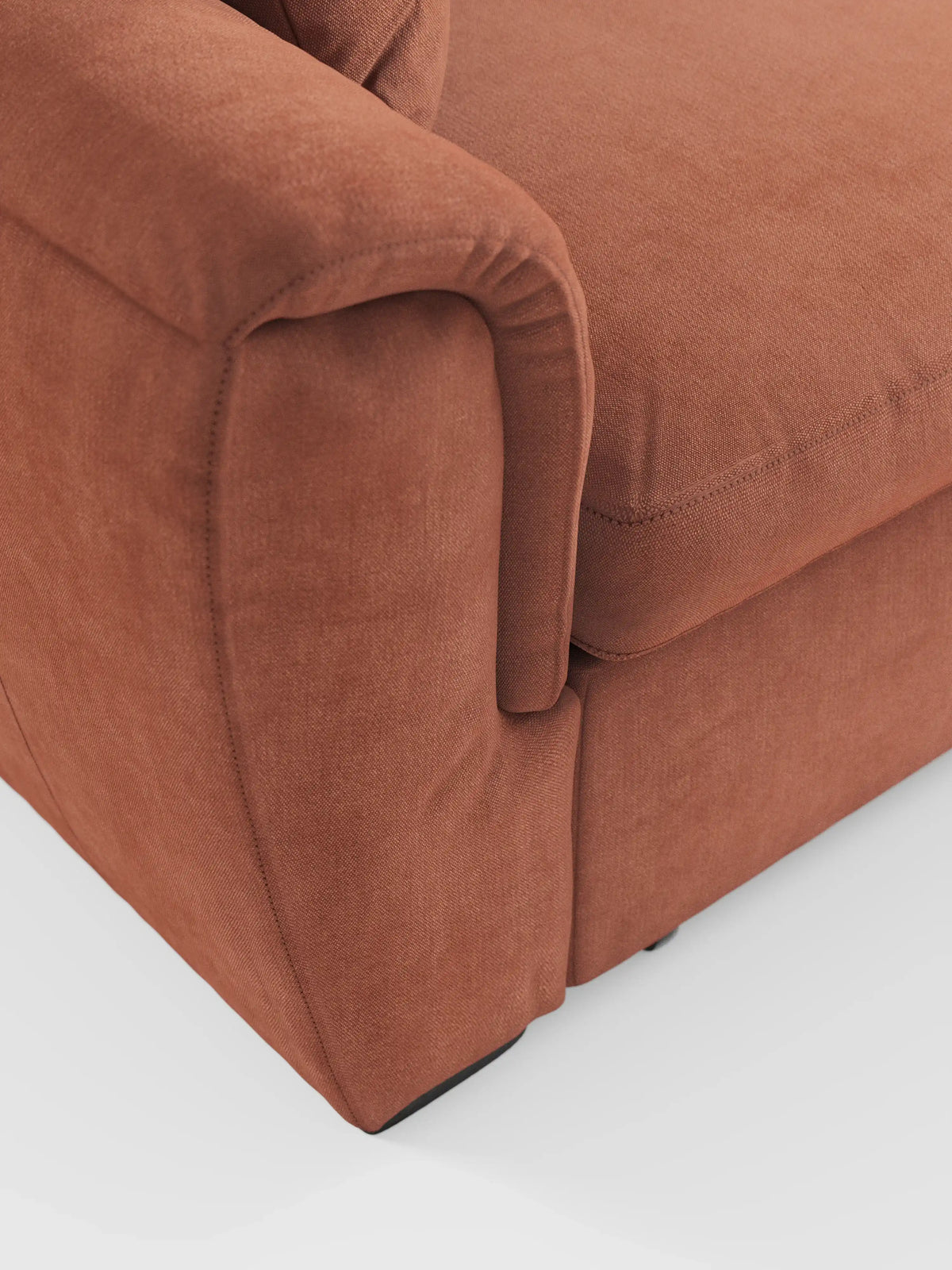 Primo piano dell'angolo anteriore del divano letto Titano con penisola reversibile e contenitore di divitt.sbs, realizzato in tessuto color ruggine e resistente alle macchie, che mostra un bracciolo, le cuciture a vista e parte del cuscino della seduta. Lo sfondo è una superficie bianca liscia.